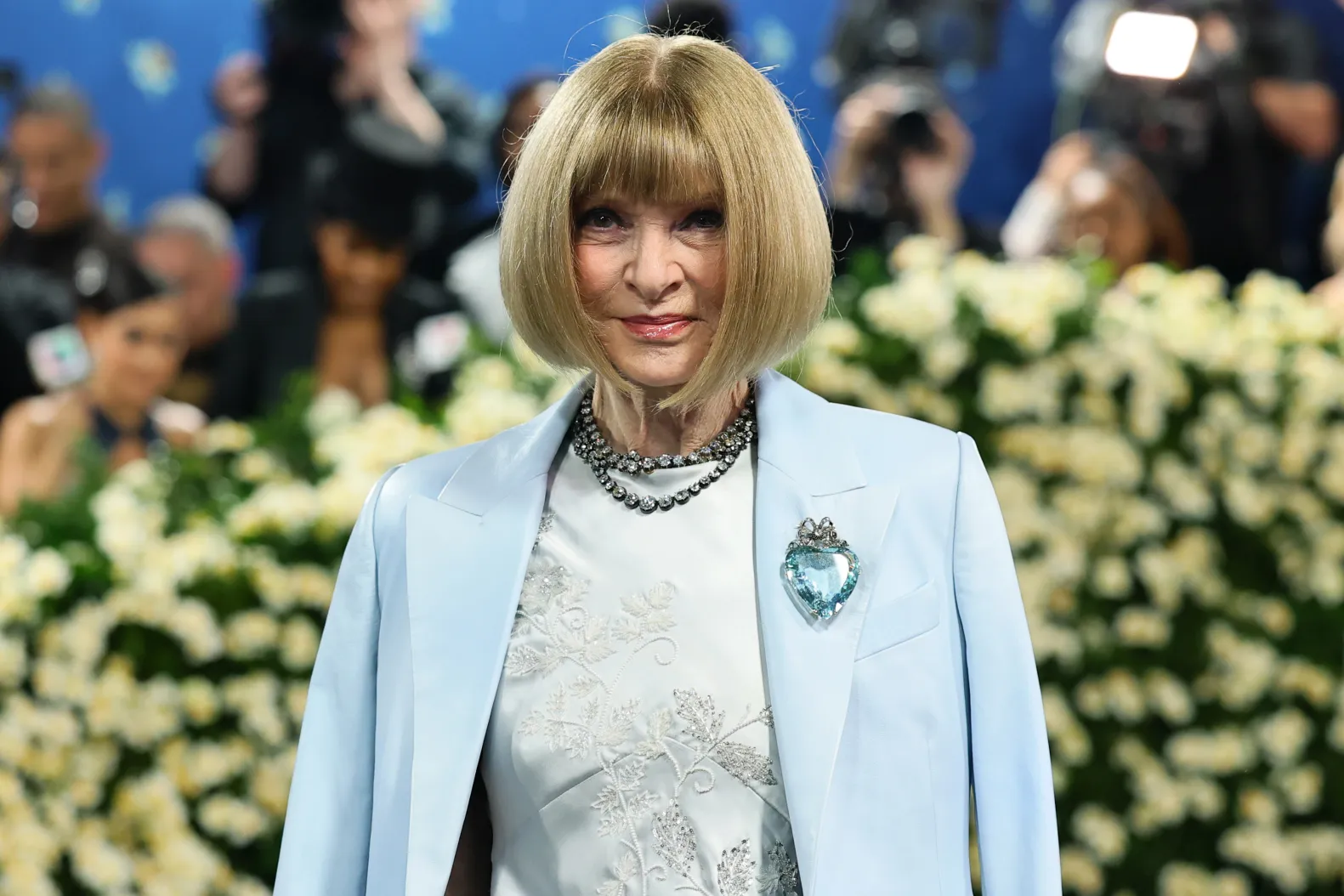 Elegant woman with a blonde bob wearing a light blue blazer and embroidered white top, accessorized with a sparkly necklace and heart-shaped brooch at a flower-filled event backdrop.