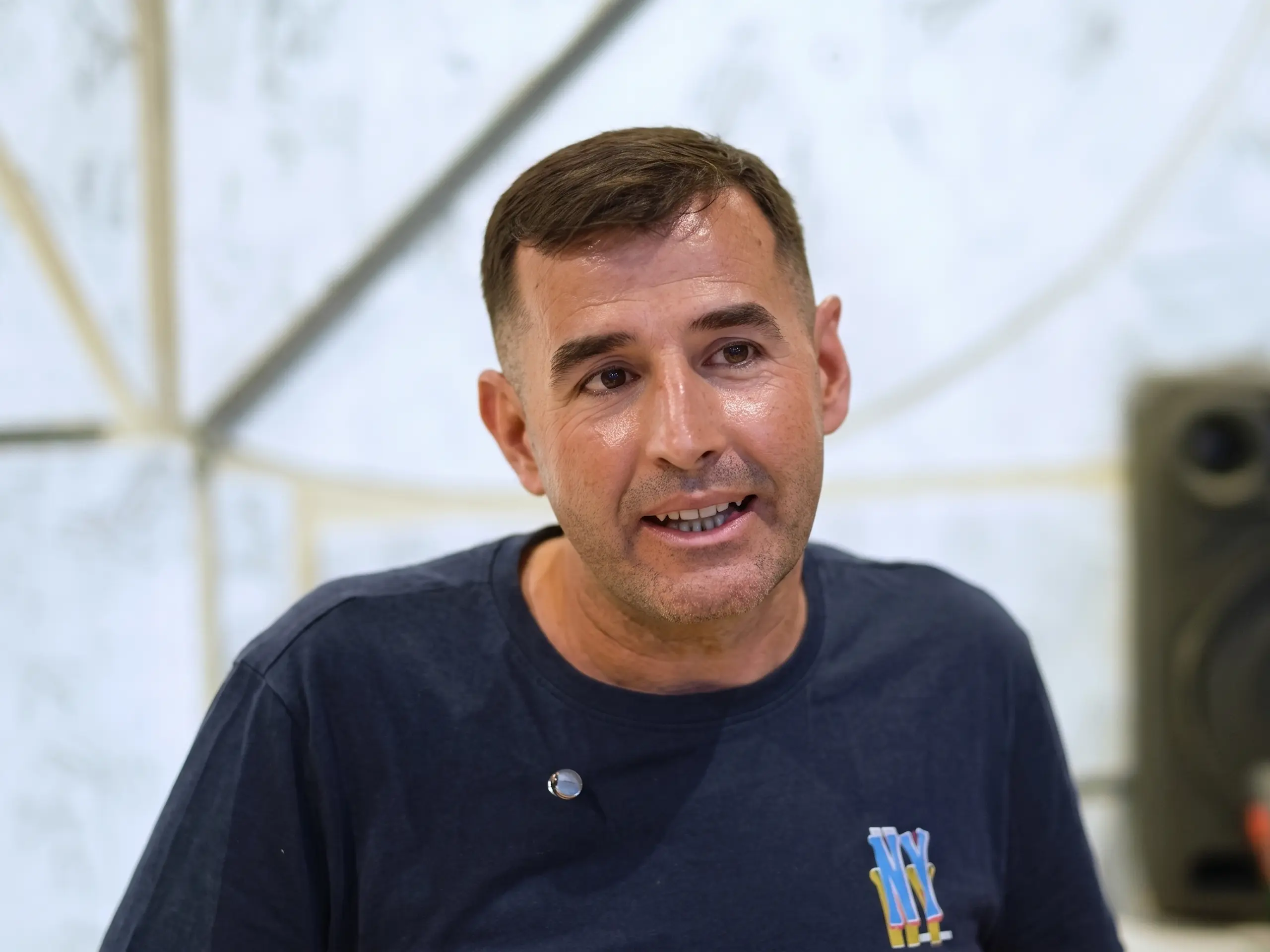 Close-up of a smiling man with short brown hair wearing a dark T-shirt, speaking in a bright indoor setting.