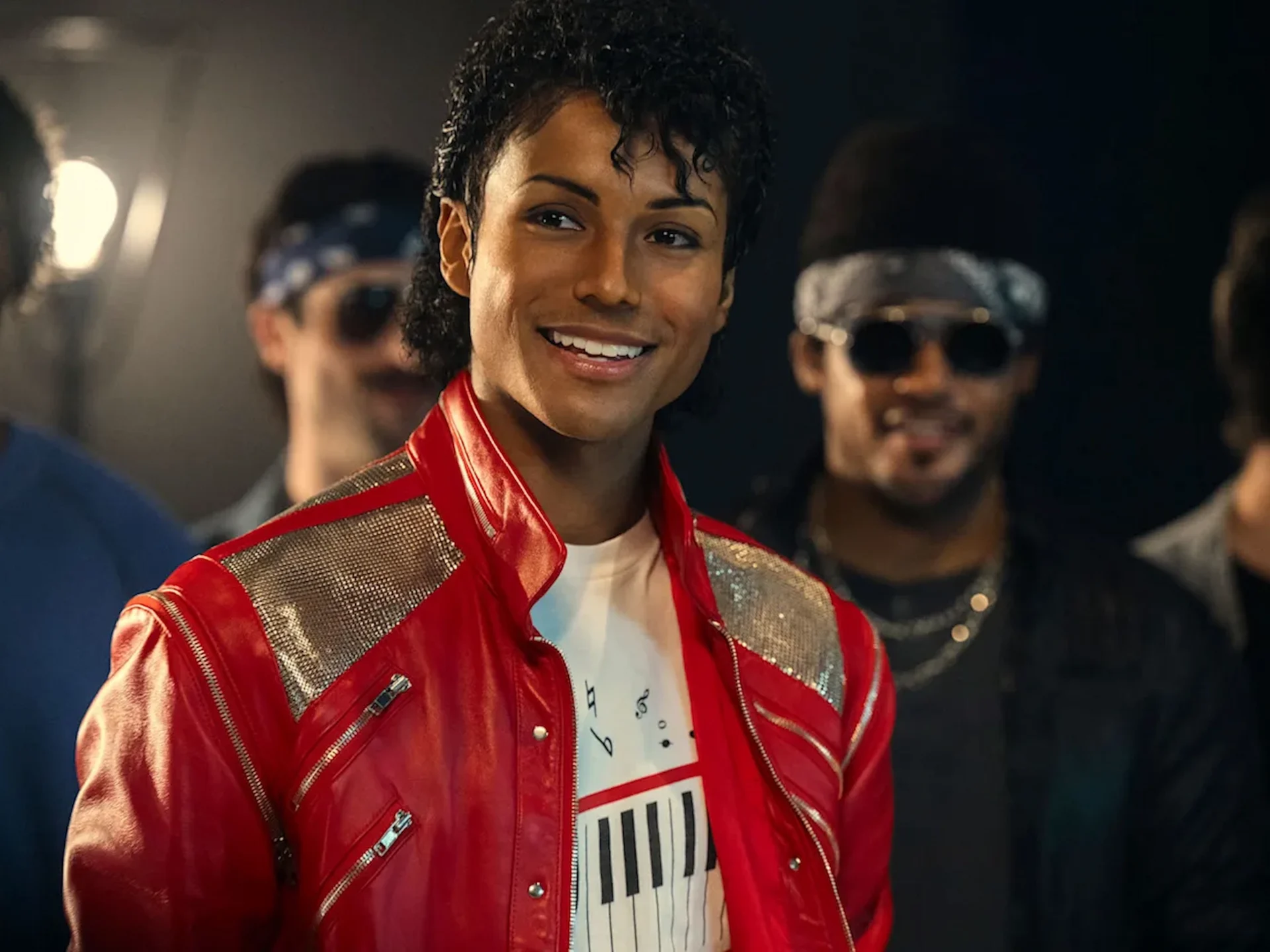 Smiling person in a shiny red jacket with silver panels, foreground, with blurred figures in a backstage setting behind them.