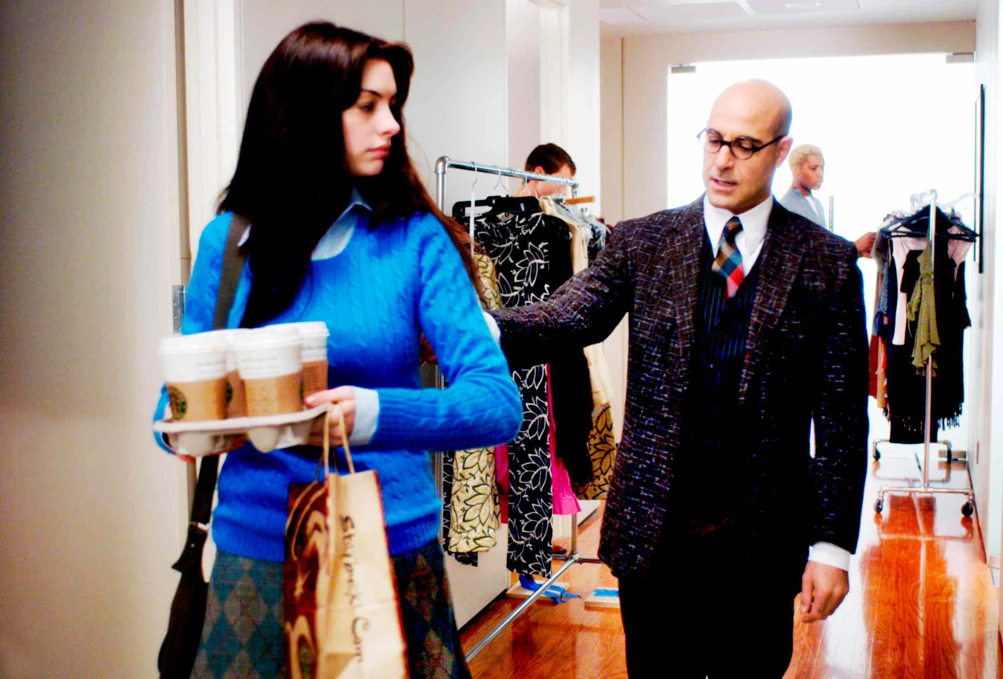 Woman in blue sweater carries coffee and shopping bags while walking past a man in a suit in a clothing store.