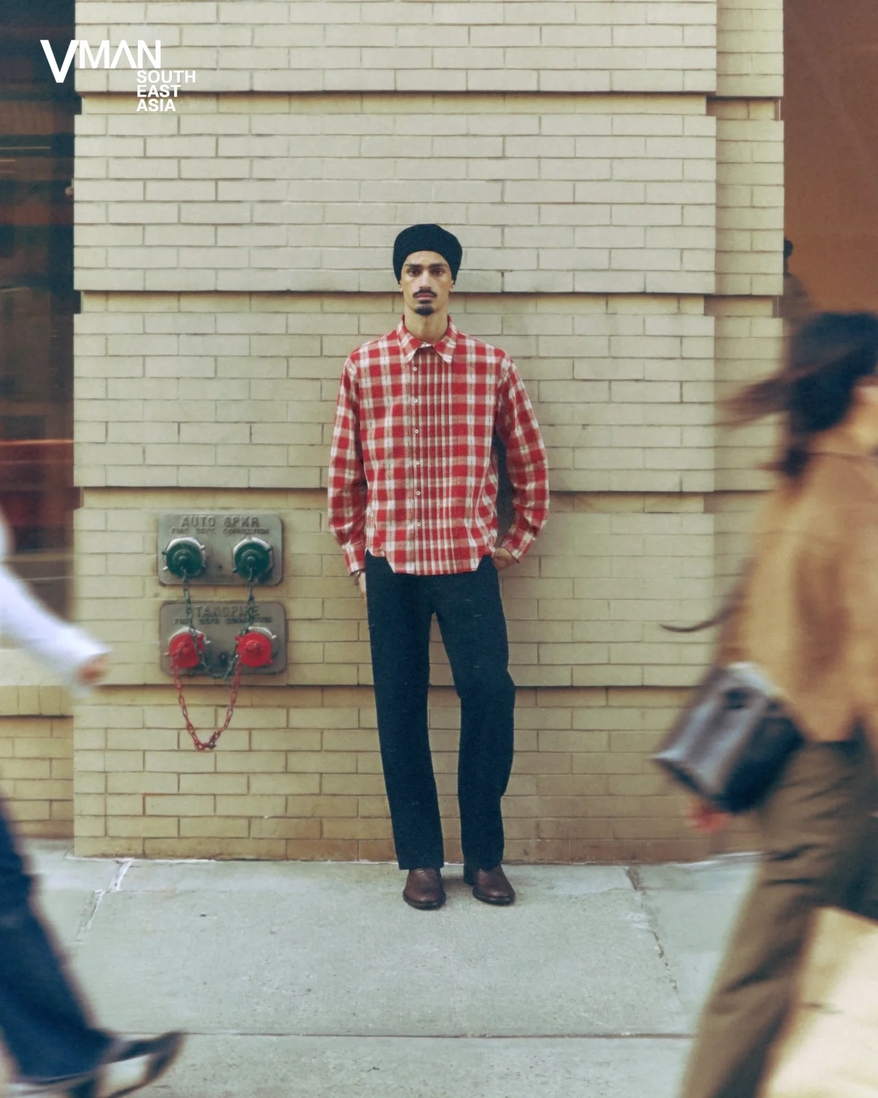 Man in a red plaid shirt and black turban stands against a beige brick wall while blurred pedestrians pass by.