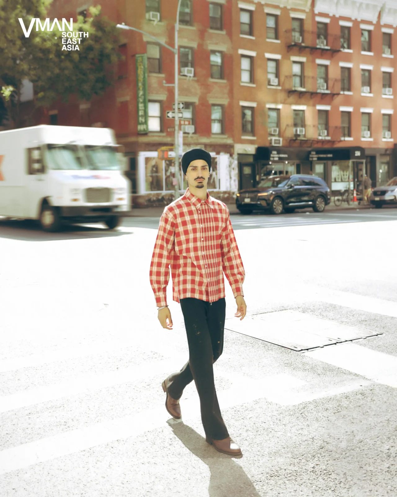 Man walks across a city crosswalk wearing a red plaid shirt and dark pants, with VMAN SE Asia logo in the corner.