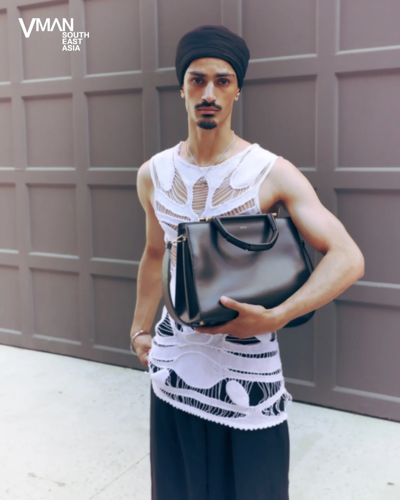 Fashion-forward man in a black turban wearing a sheer white crochet tank top, holding a black handbag against a gray panel wall.