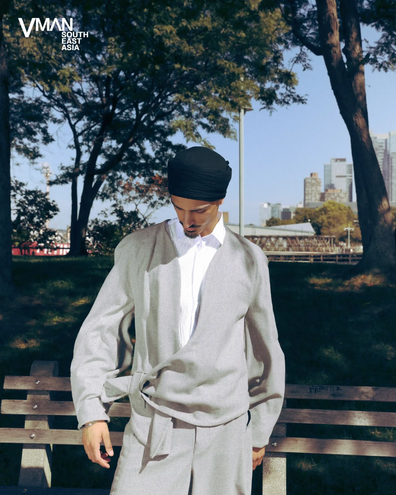 Man in a light gray robe with white shirt and black turban stands beside a wooden bench in a sunny urban park, city skyline in the background.