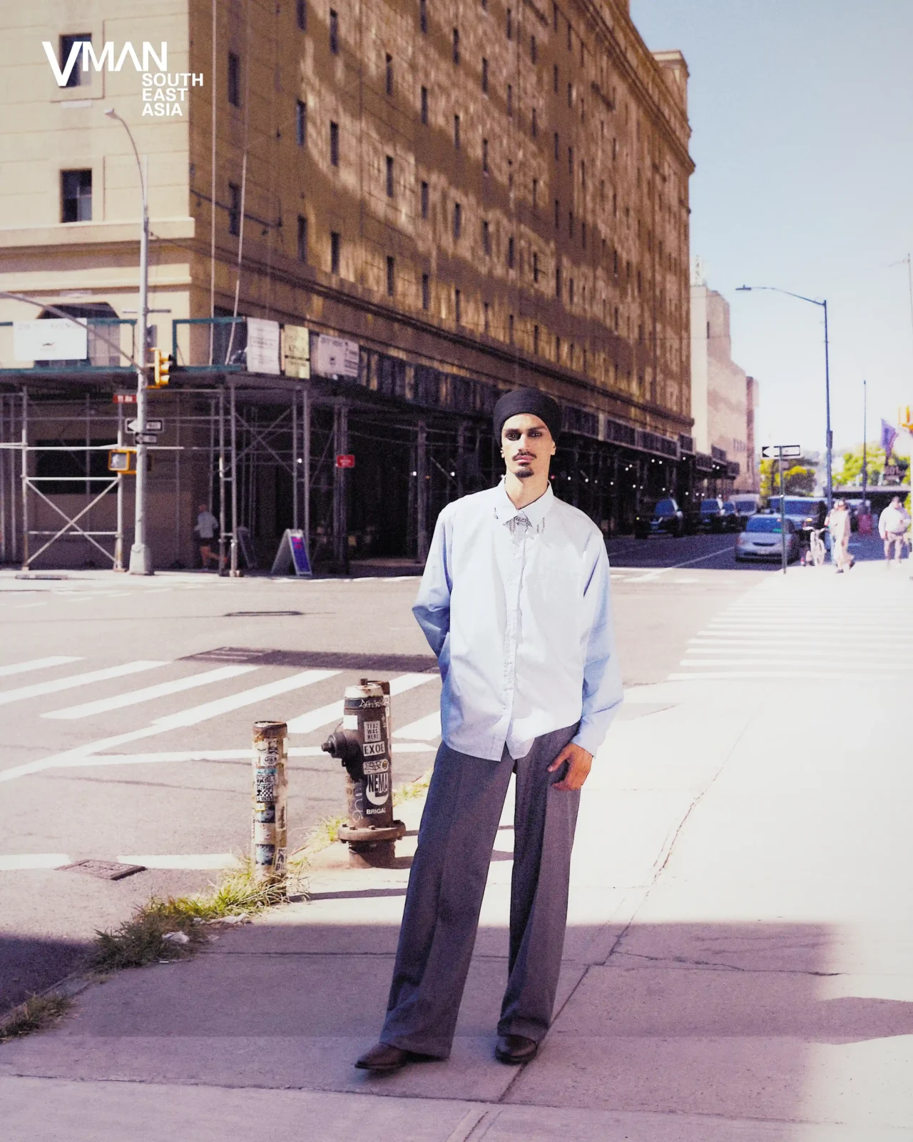Male model in a pale blue shirt and wide gray trousers stands on a city sidewalk at a crosswalk, with a tall beige building behind and VMAN SE Asia logo in the corner.