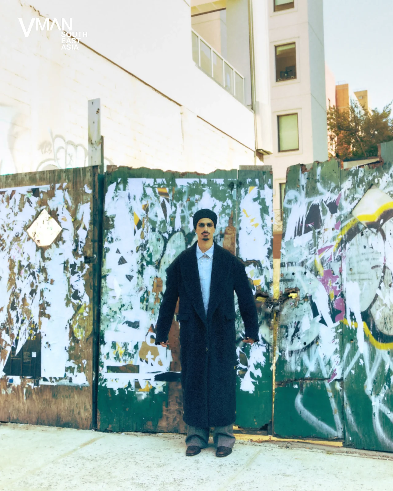 Man wearing a long dark overcoat stands in front of a graffiti-covered fence in an urban setting, looking at the camera.