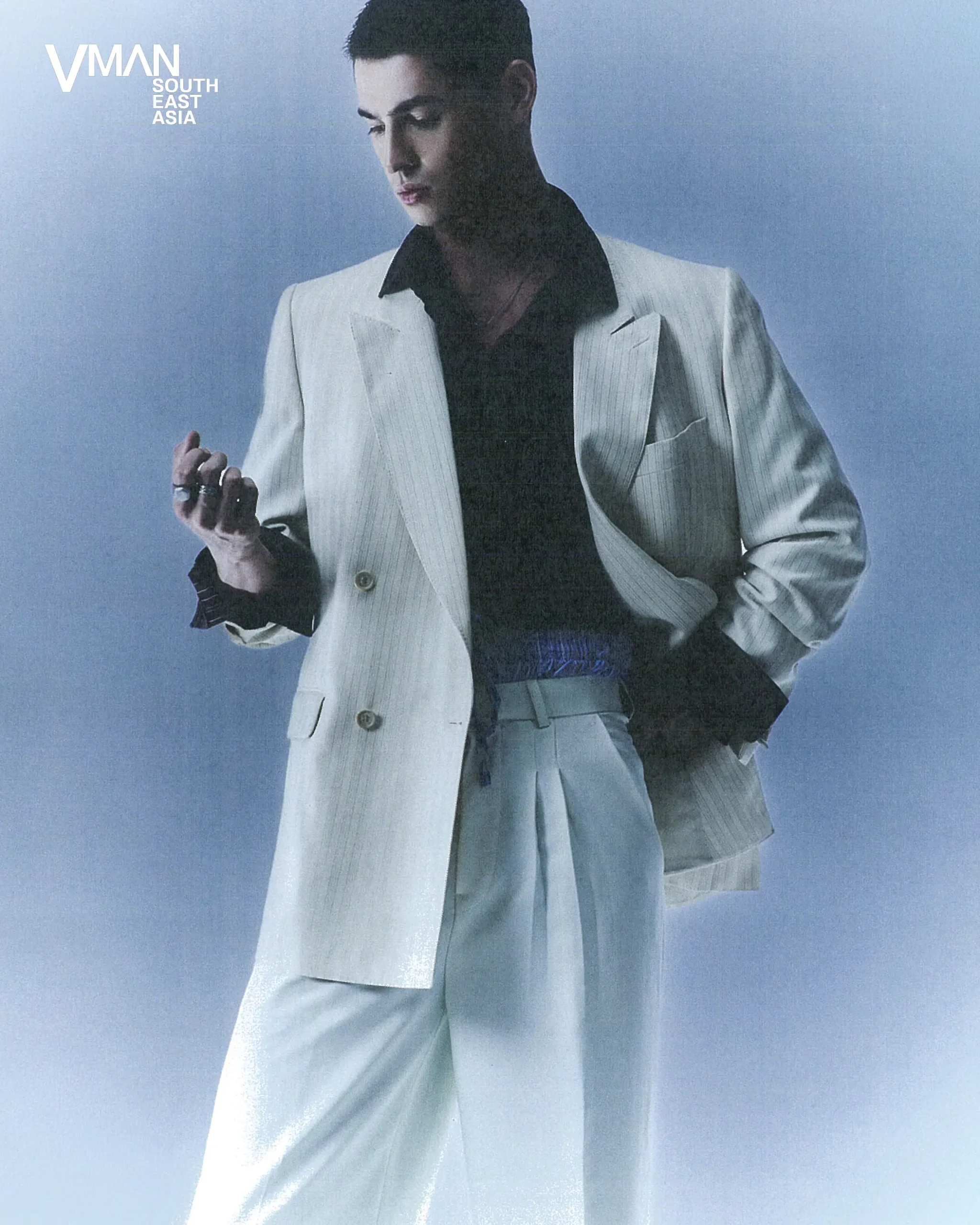 Male model in a light pinstripe suit and black shirt poses against a blue studio backdrop with the VMAN South East Asia logo in the corner.