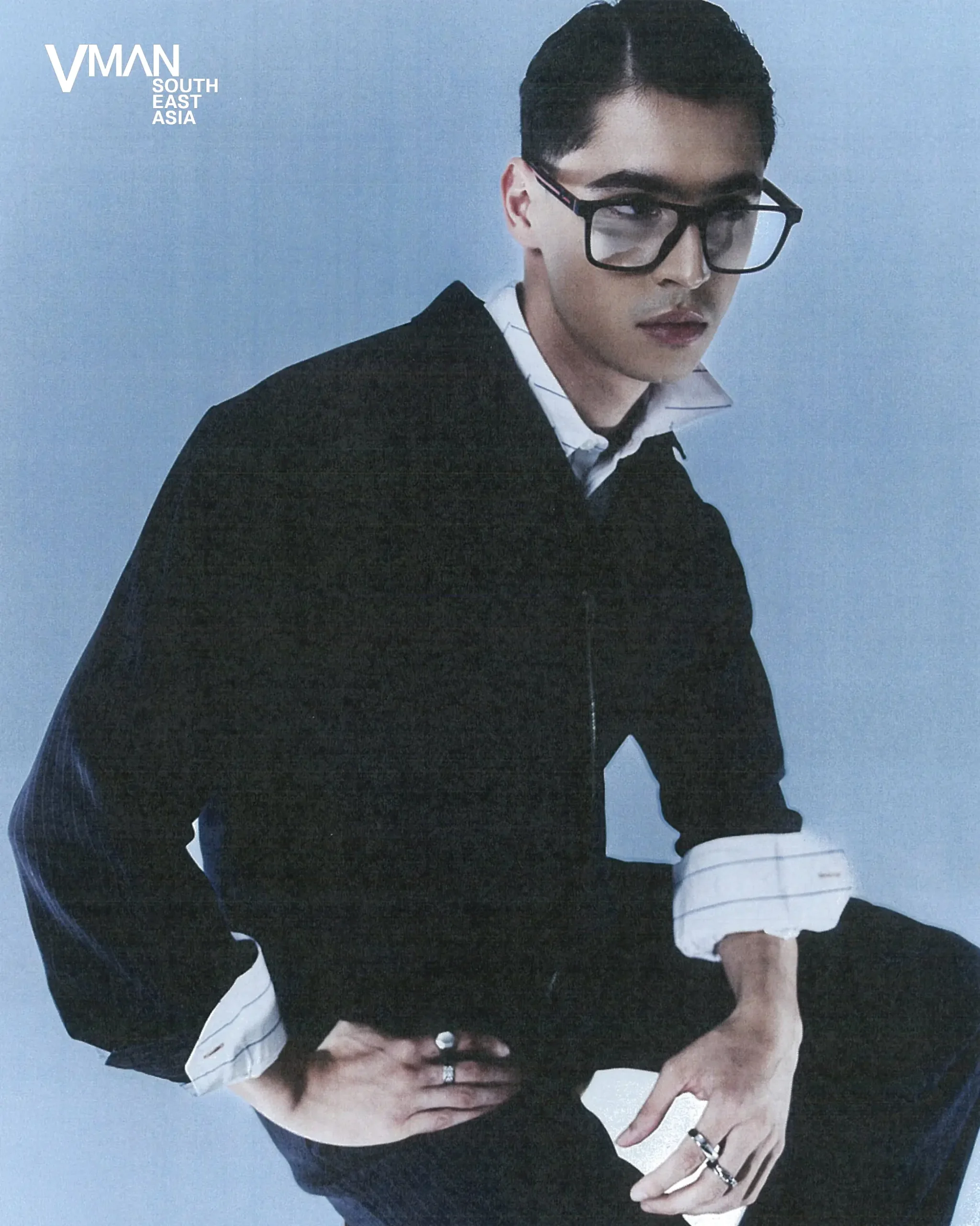 Man posing in a black sweater with a white shirt, oversized glasses, against a blue backdrop; VMAN South East Asia logo in the corner.