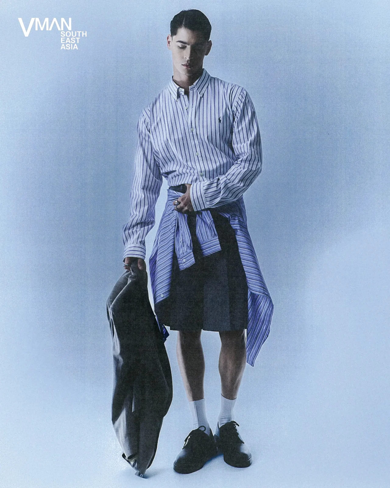Man wearing a blue striped button-down shirt with a wrap-around jacket, standing against a pale blue studio backdrop.