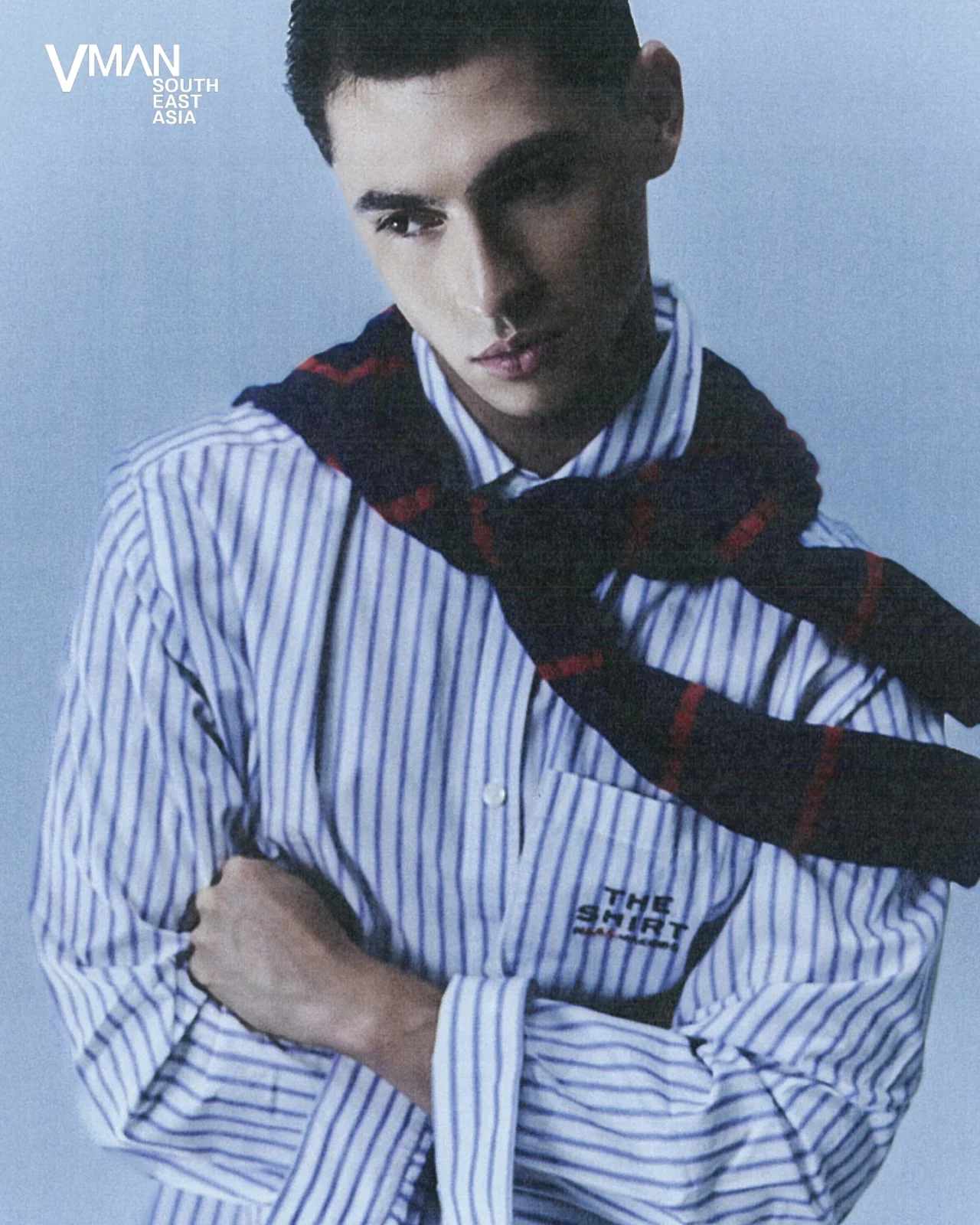 Male model in a blue-and-white striped shirt with a dark sweater draped over his shoulders, posed against a blue studio backdrop with the VMAN South East Asia logo in the corner.