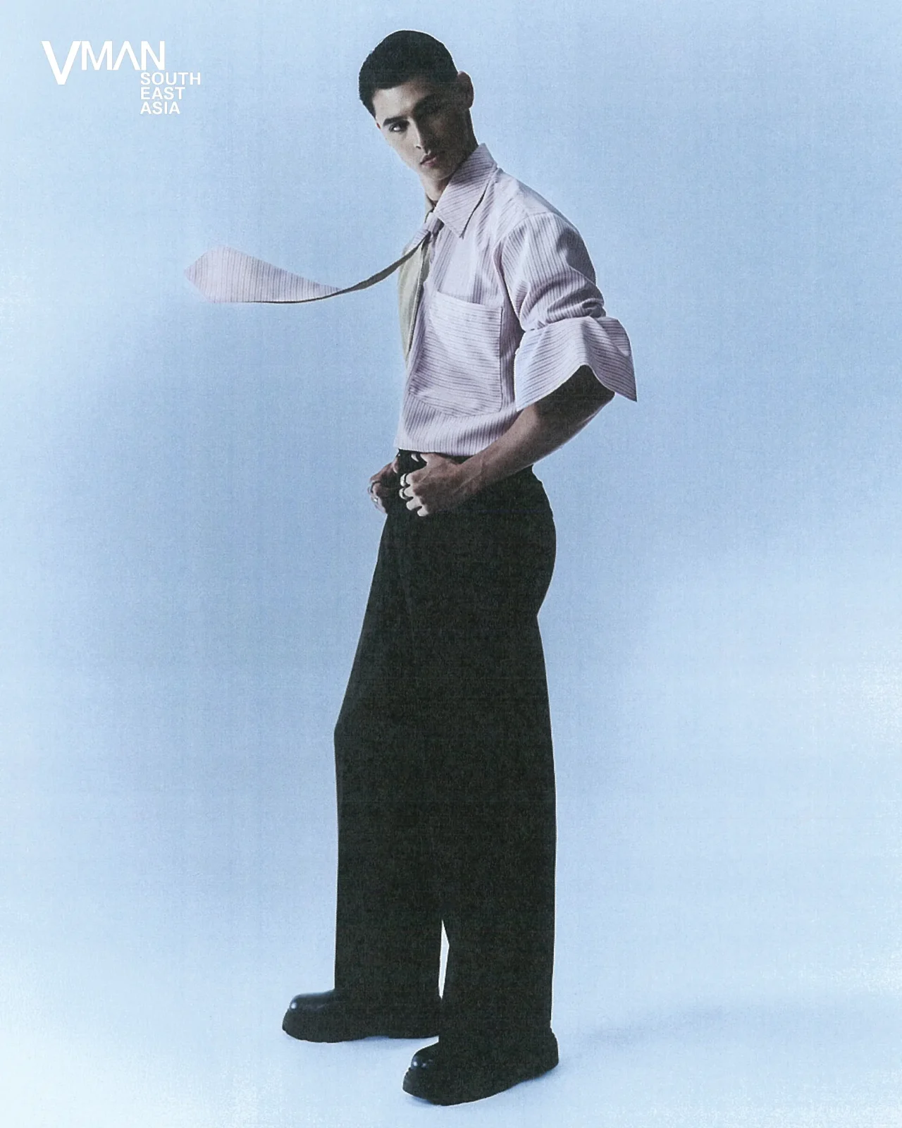 Model standing in a light striped shirt with a loose tie, hands on hips, against a pale blue backdrop; VMAN South East Asia logo visible in the corner.