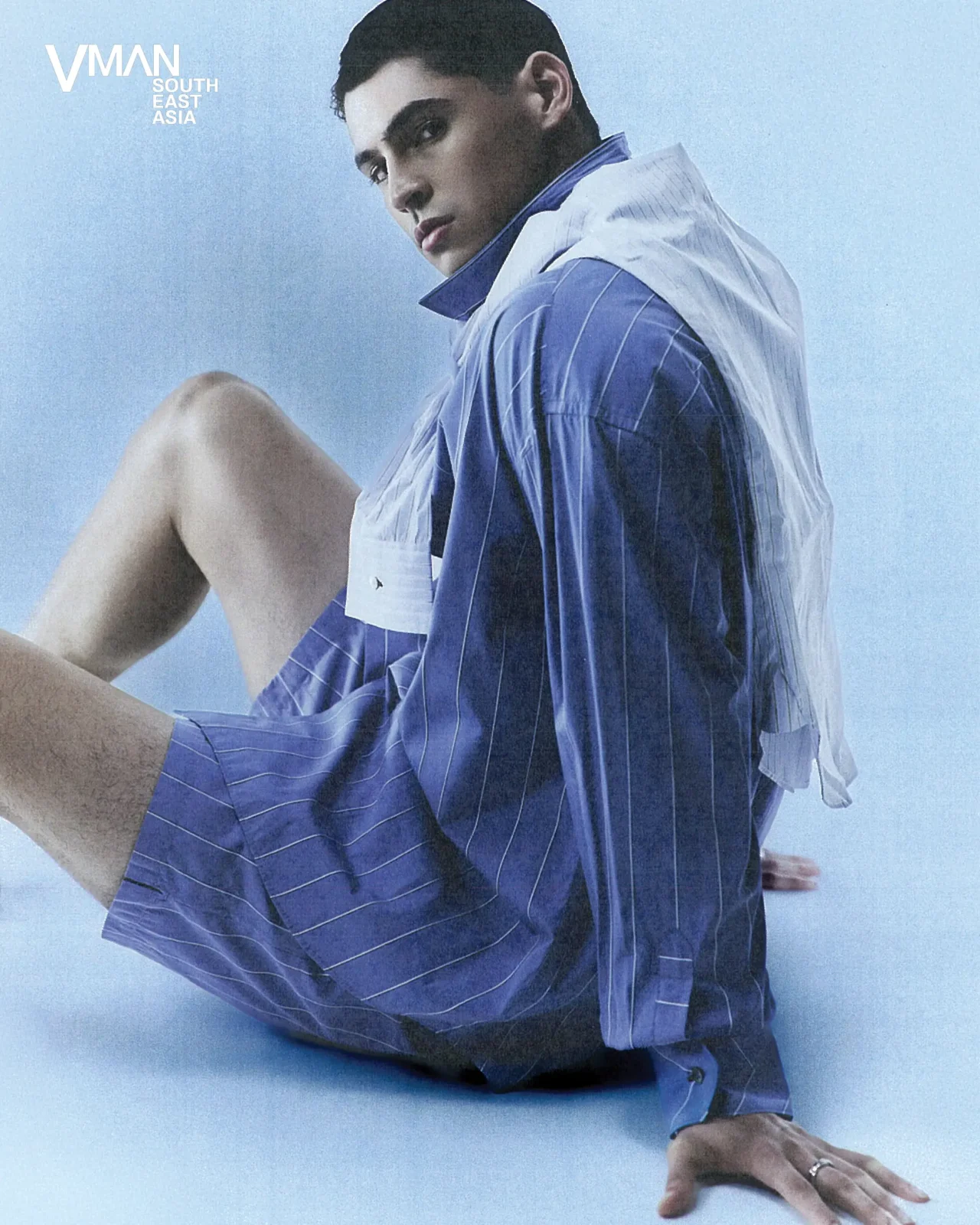 Model seated in a blue pinstripe shirt against a pale blue background; VMAN South East Asia logo upper left.