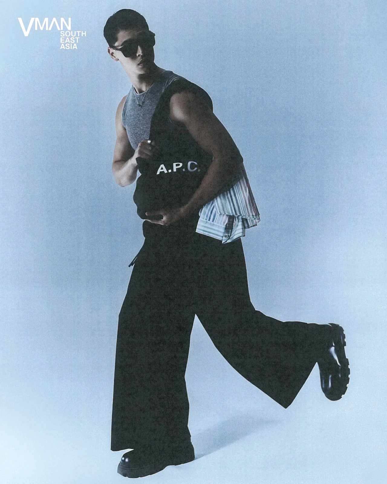 Male model running in a gray sleeveless top with 'A.P.C.' on it, black pants, sunglasses, and a striped shirt tied around the waist against a blue backdrop; VMAN South East Asia logo visible in the corner.