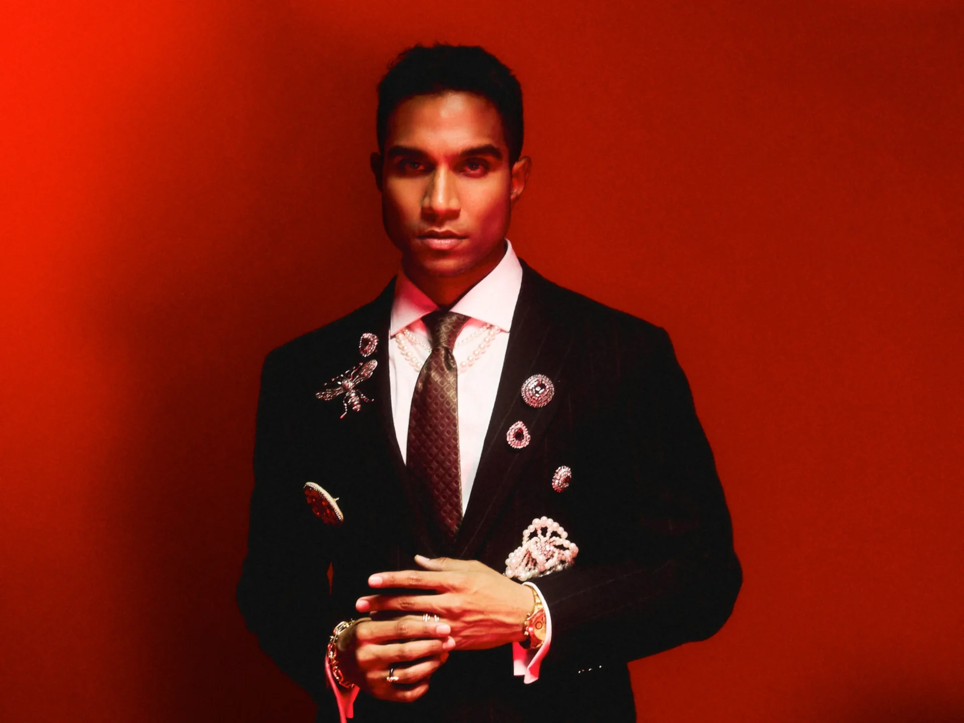 Portrait of a person in a dark suit with lapel pins, white shirt, and brown patterned tie against a vivid red backdrop.