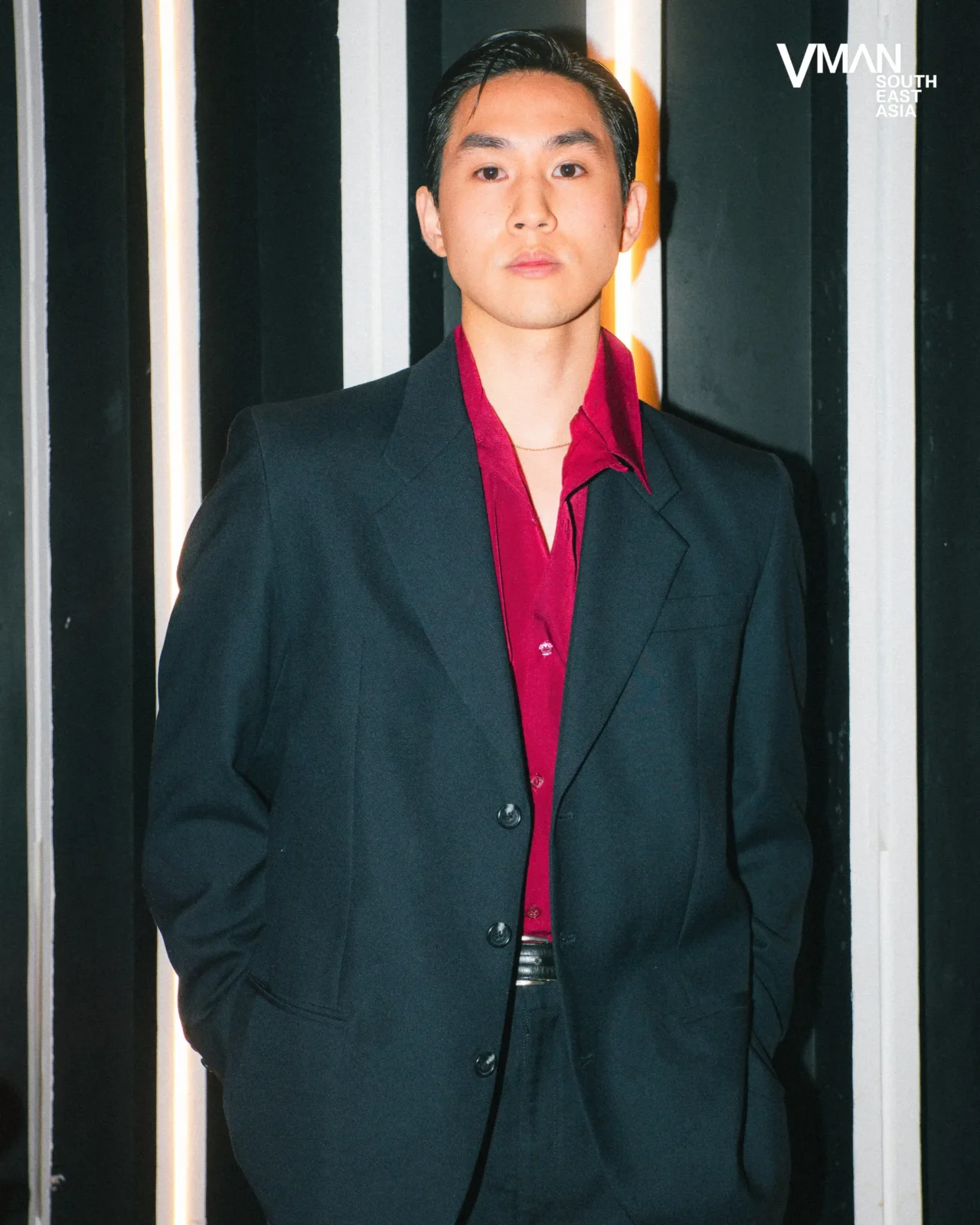 Young man in a black blazer and red shirt, hands in pockets, standing against a striped backdrop.