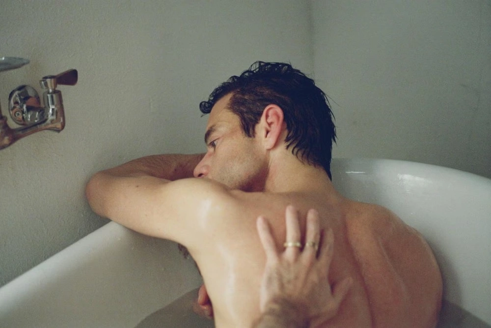 Man lying in a bathtub face turned away, resting his head on the edge while another hand touches his back gently.