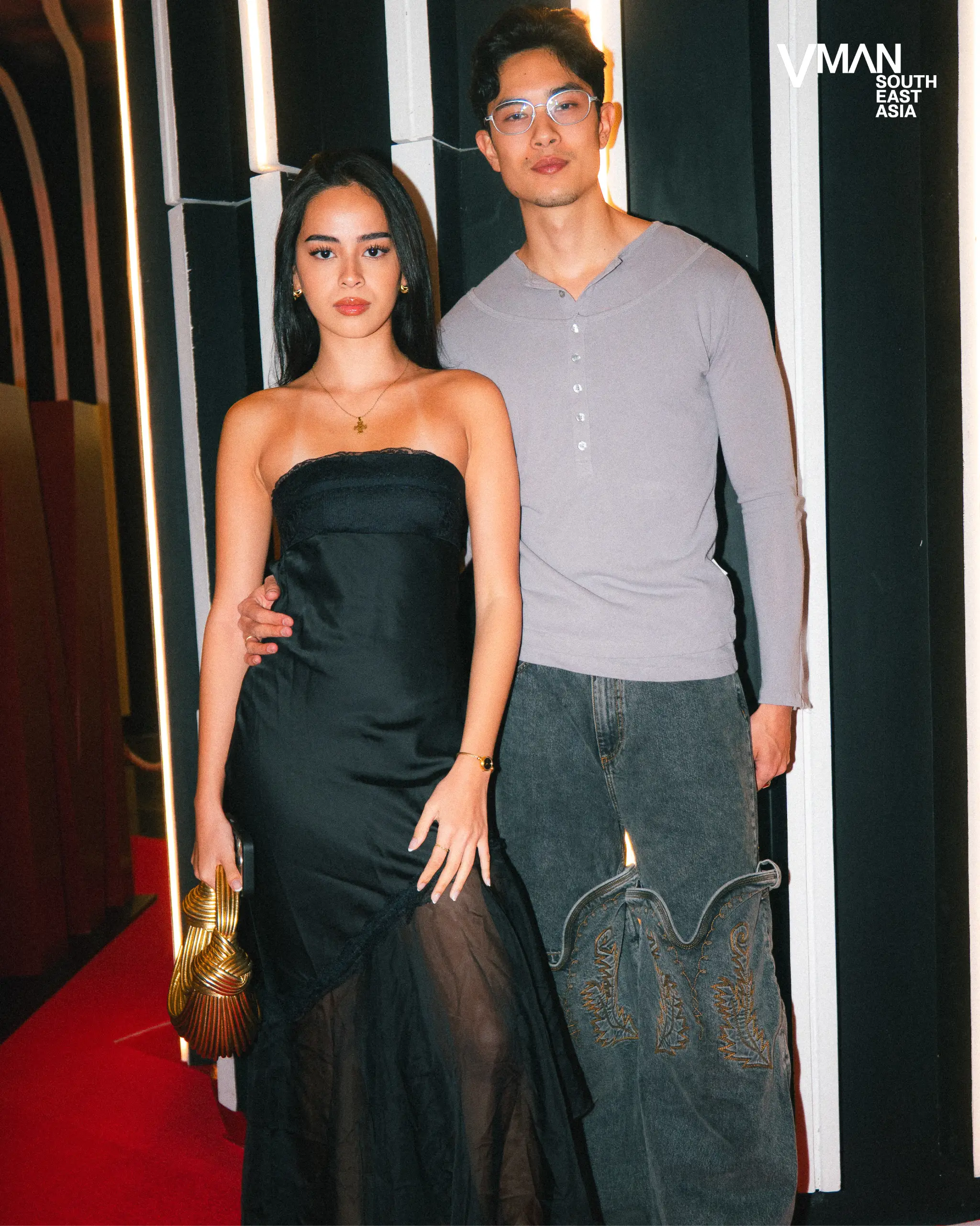 Two stylish models pose on a red carpet with a VMAN South East Asia backdrop; woman in a black strapless dress holding a gold clutch, man in a gray long-sleeve shirt and jeans beside her.