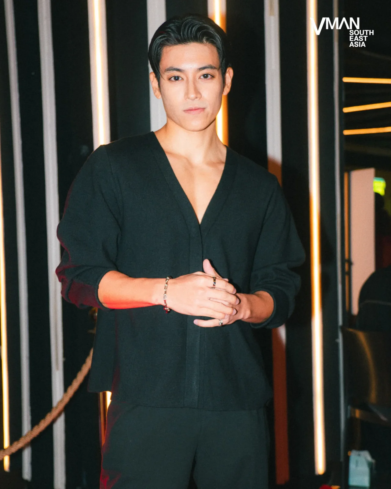 Man wearing a black V-neck top stands with hands clasped in front of vertical light bars, VMAN South East Asia logo in the background.