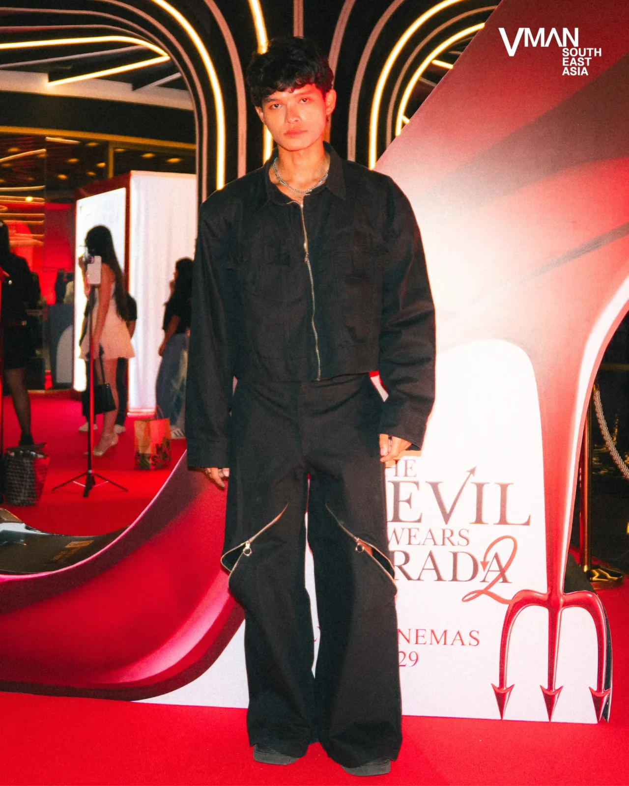 Young man in a black zipped jacket and chain necklace posing on a red carpet with neon arch lights behind him.