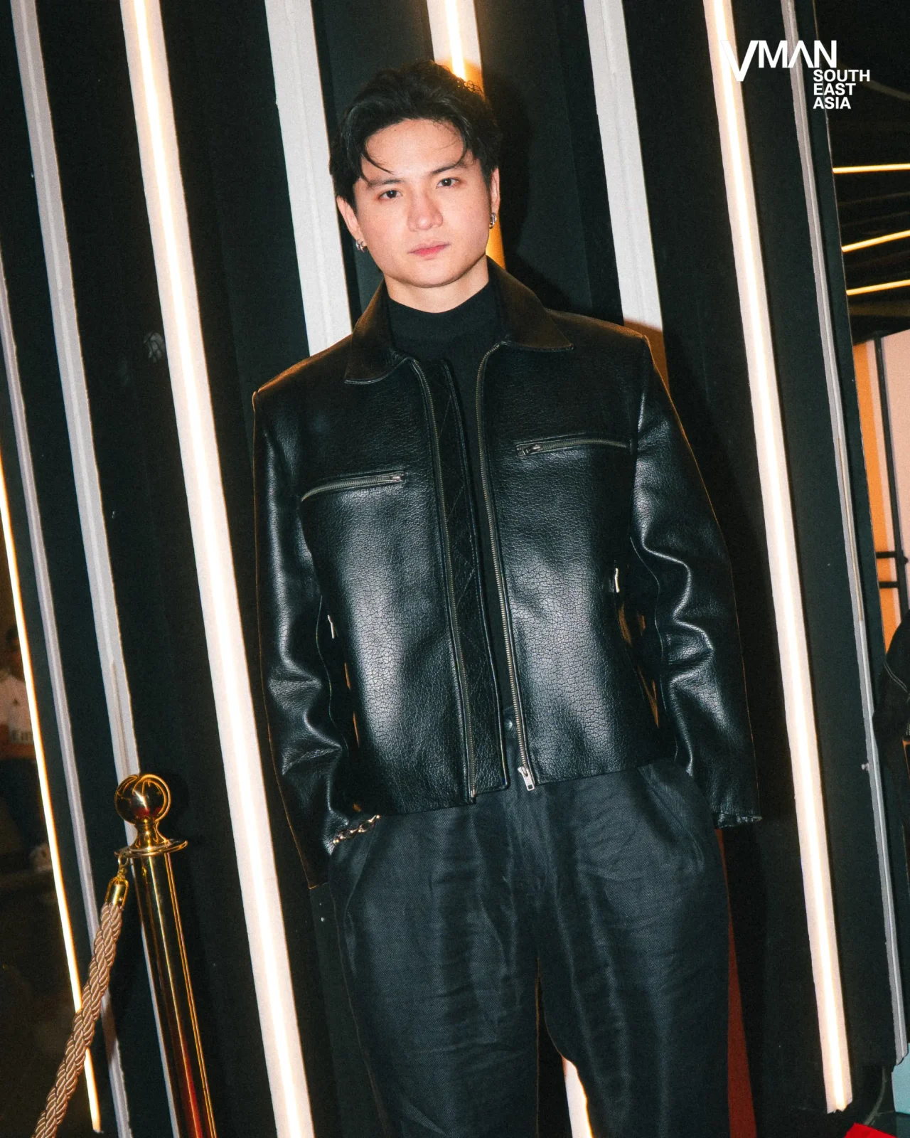Mannequin wearing a black leather jacket and turtleneck against a striped wall with vertical lights; VMAN South East Asia logo in corner.