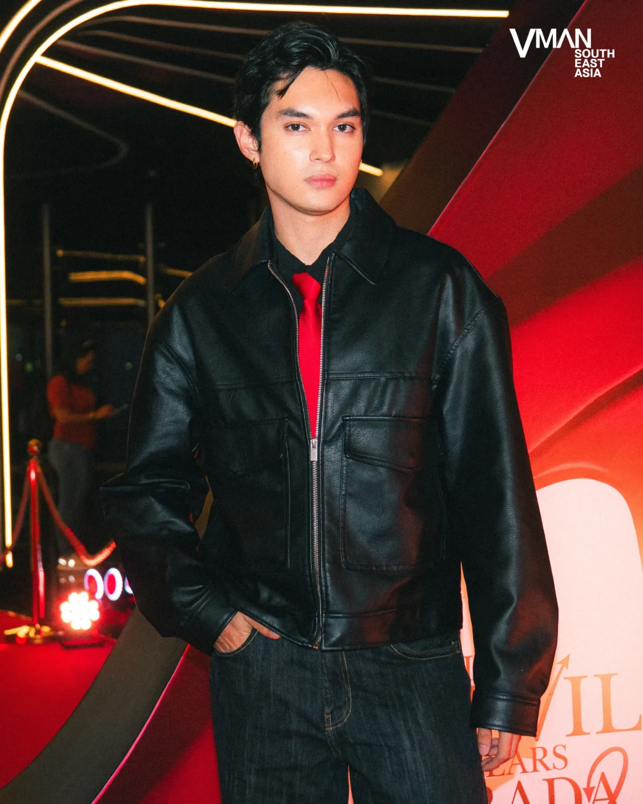 Young man in a black leather jacket and red tie posing indoors against neon lights and a VMAN Southeast Asia logo backdrop.