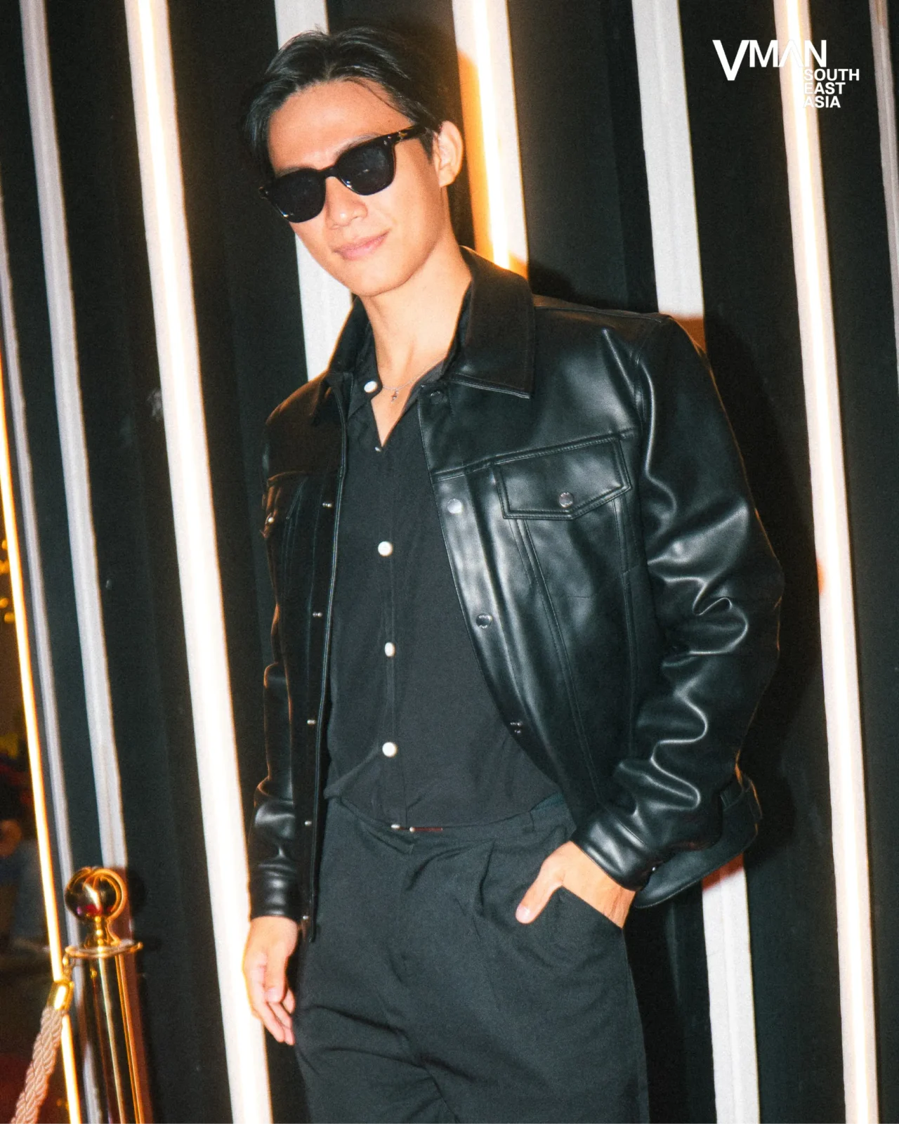 Man in a black leather jacket and sunglasses posing against striped black-and-white backdrop with a logo in the corner (VMAN Southeast Asia).