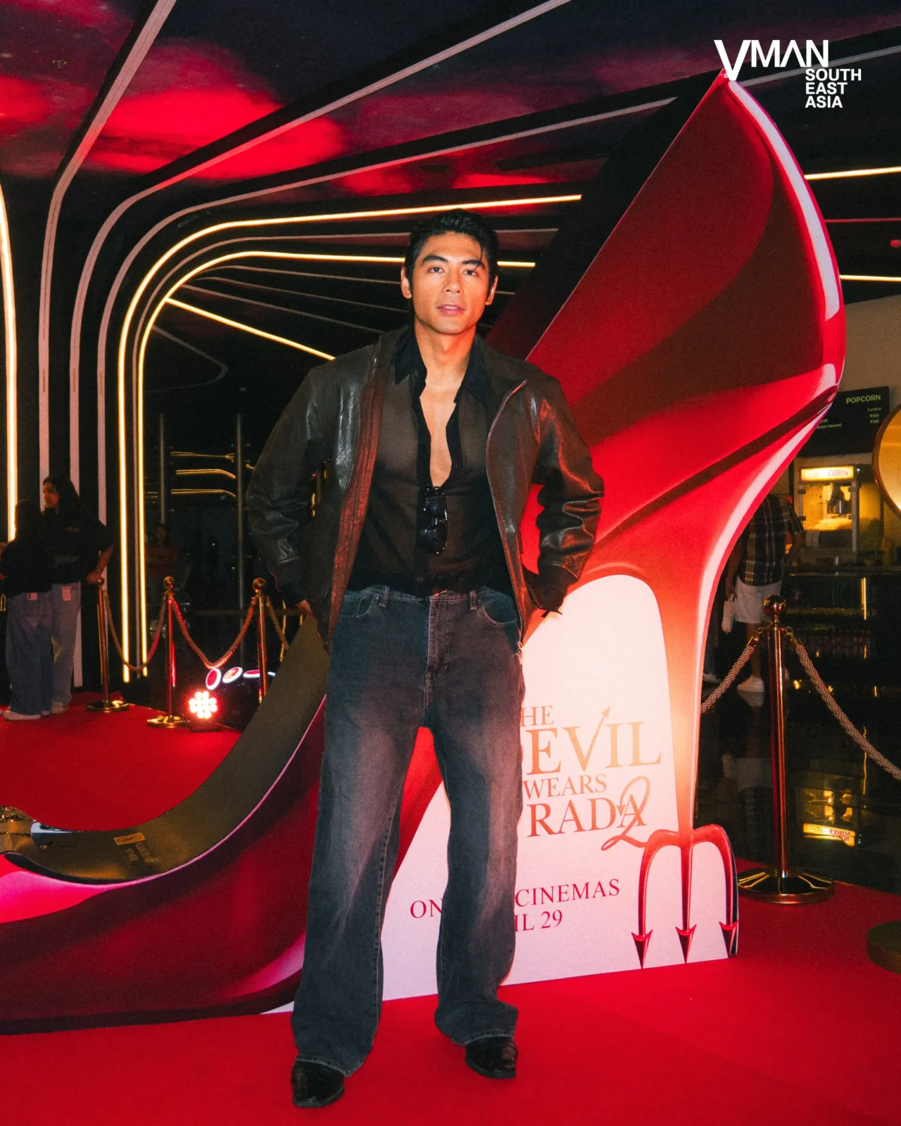 Man in a black leather jacket and dark jeans poses on a red carpet with neon curved arches behind him at a film event.
