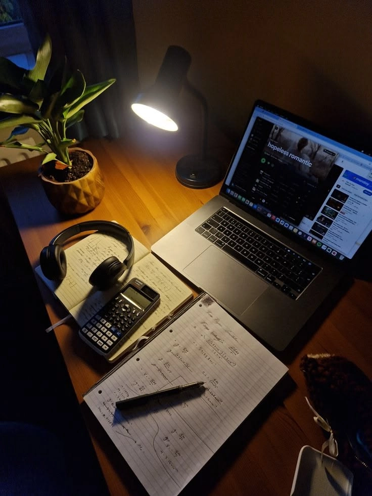 Nighttime desk setup with a laptop, glowing lamp, potted plant, headphones, calculator, and a notebook with handwritten notes.