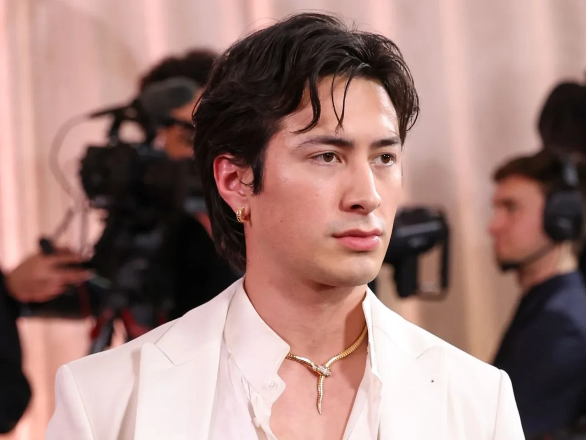 Close-up of a man with black hair and a gold hoop earring wearing a white suit at a red-carpet event, photographers in the background.