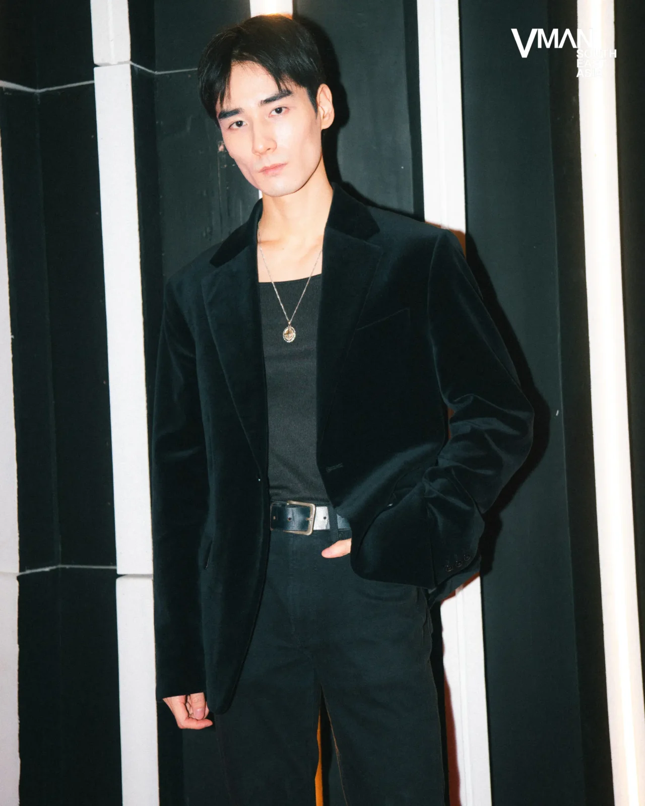 Man in a dark velvet blazer and black shirt stands against a black-and-white striped wall, wearing a pendant necklace.
