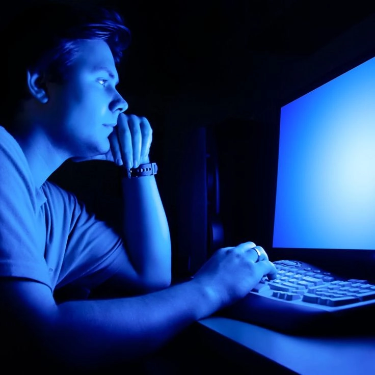Young man in a dark room lit by a blue computer screen, resting chin on his hand and typing on the keyboard.