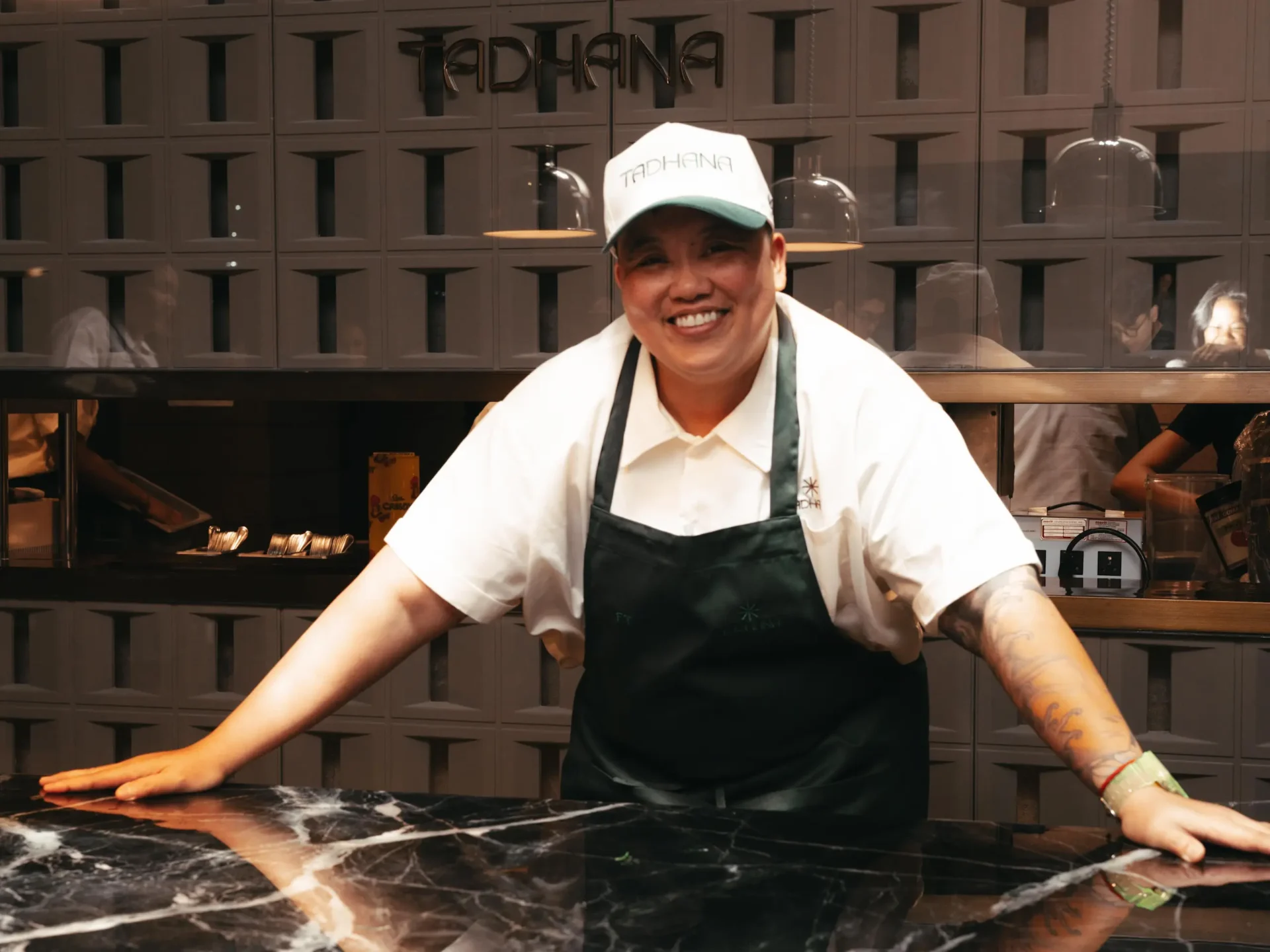 Smiling chef in a white shirt and dark apron leaning over a marble counter in a professional kitchen, with a tile wall behind.