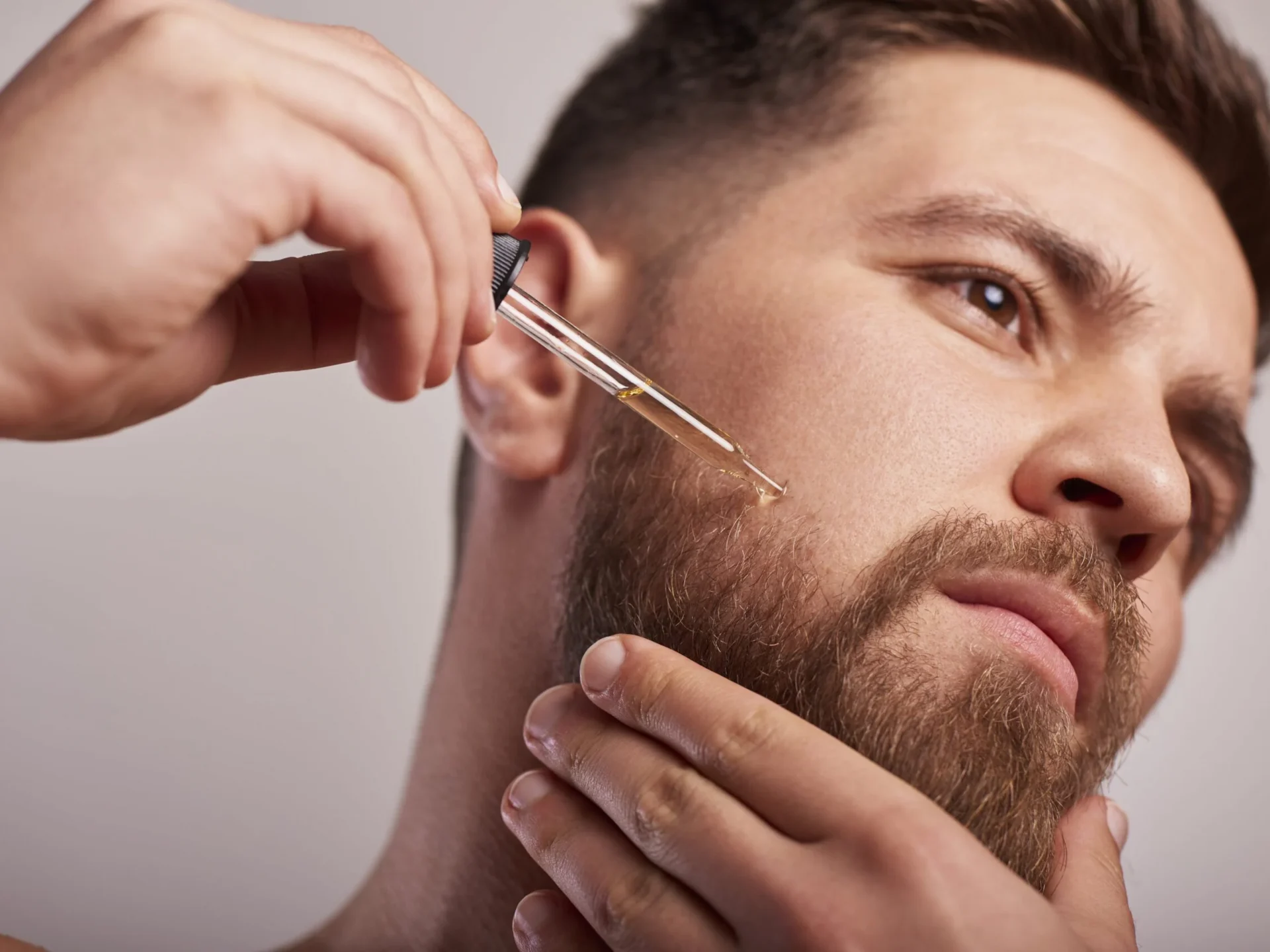 Close-up of a man applying beard oil with a dropper to his beard area near the jawline