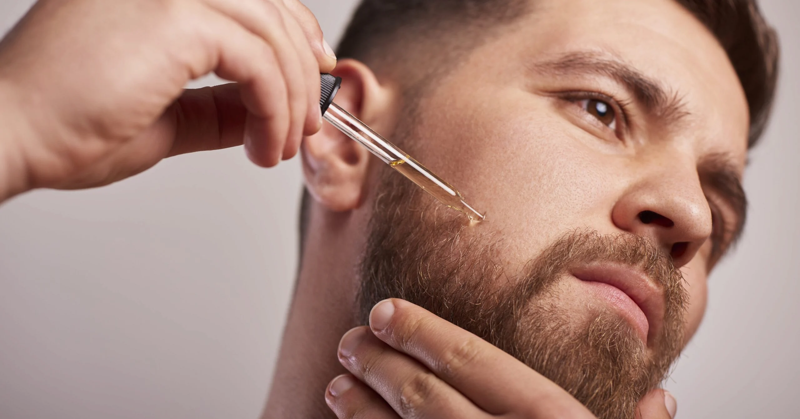 Close-up of a man applying beard oil with a dropper to his beard area near the jawline