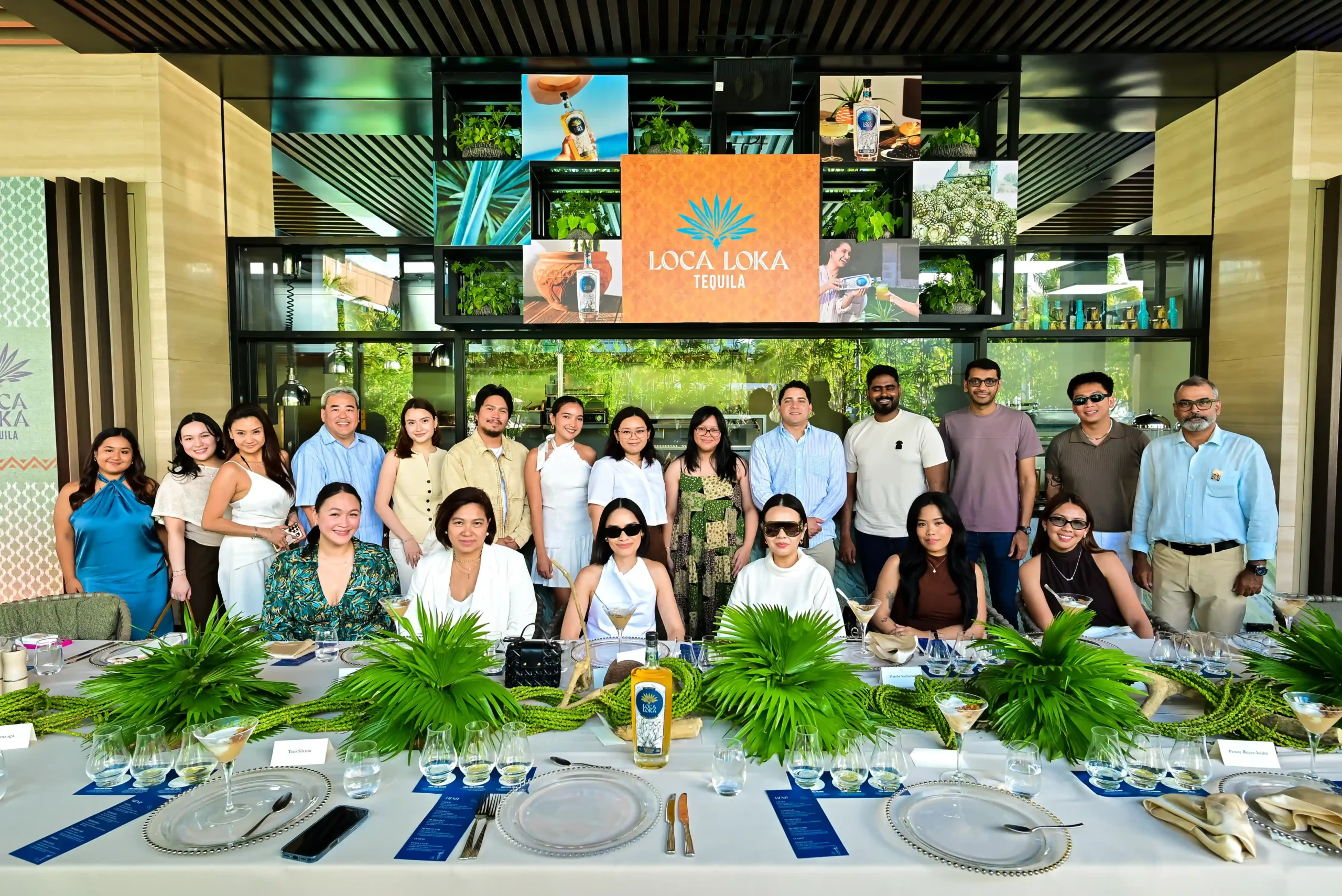 Group of people standing in a banquet setting, with a long, neatly set table and a Loca Loka Tequila display behind them.