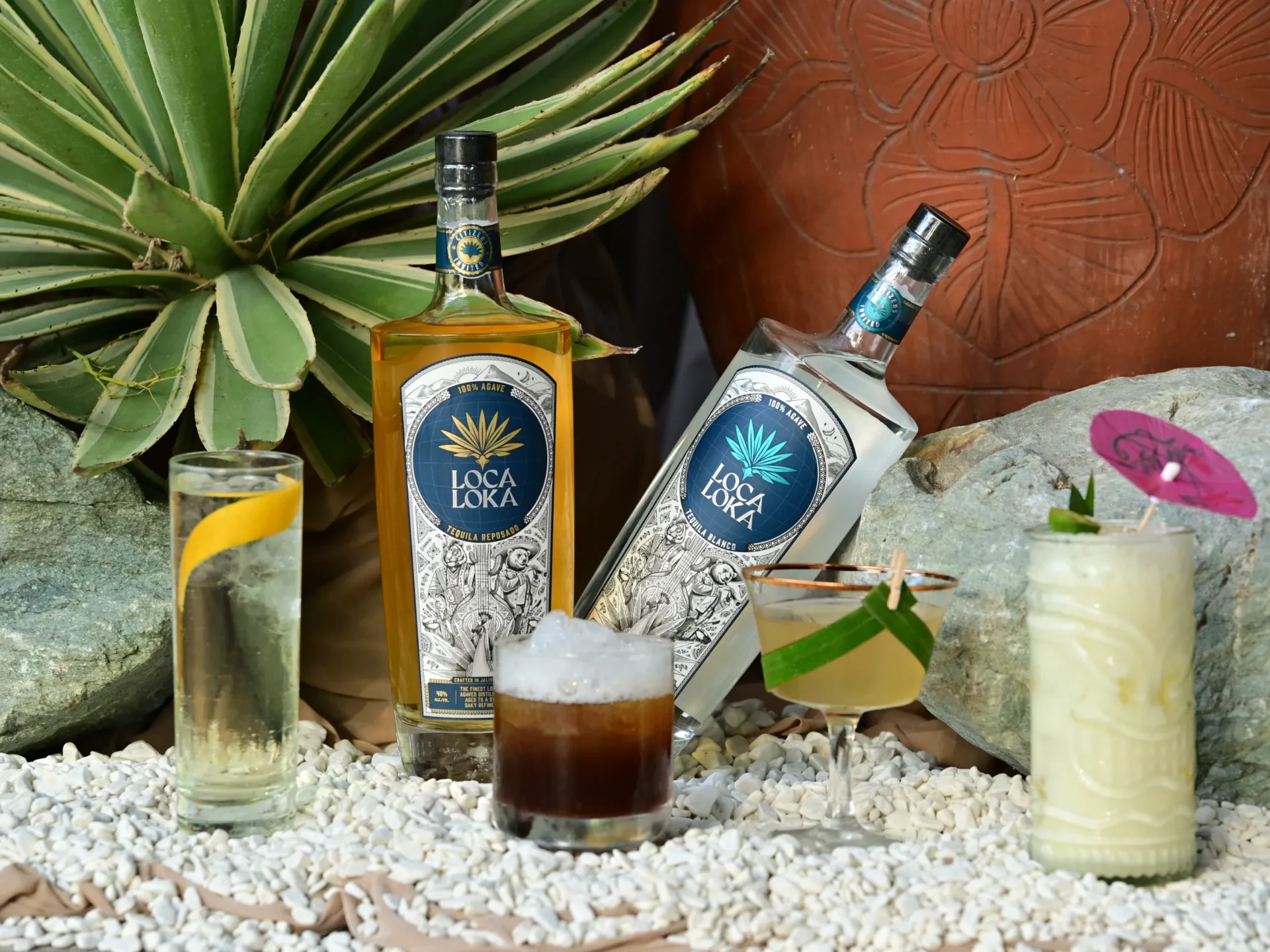 Two Loca Loka tequila bottles (reposado and blanco) with cocktails and tropical plants in a rocky, outdoor setting