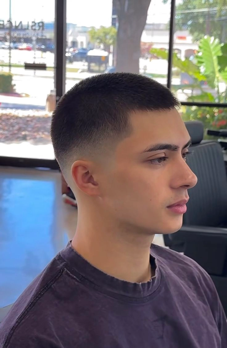 Side profile of a young man with a clean high fade haircut sitting in a barber chair inside a sunny salon.