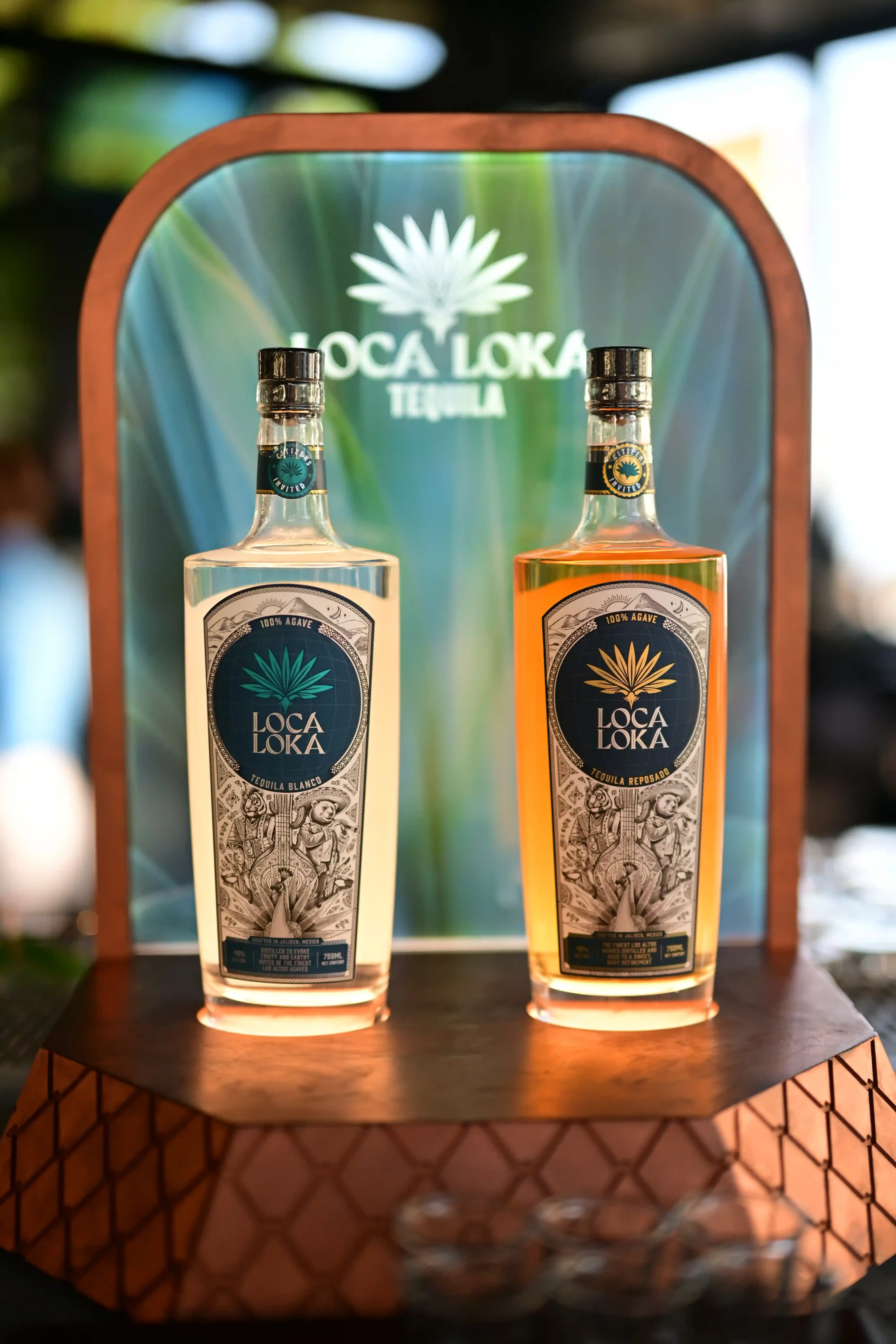 Two Loca Loka tequila bottles on a round wooden counter in front of a teal display backdrop with the brand logo behind them.