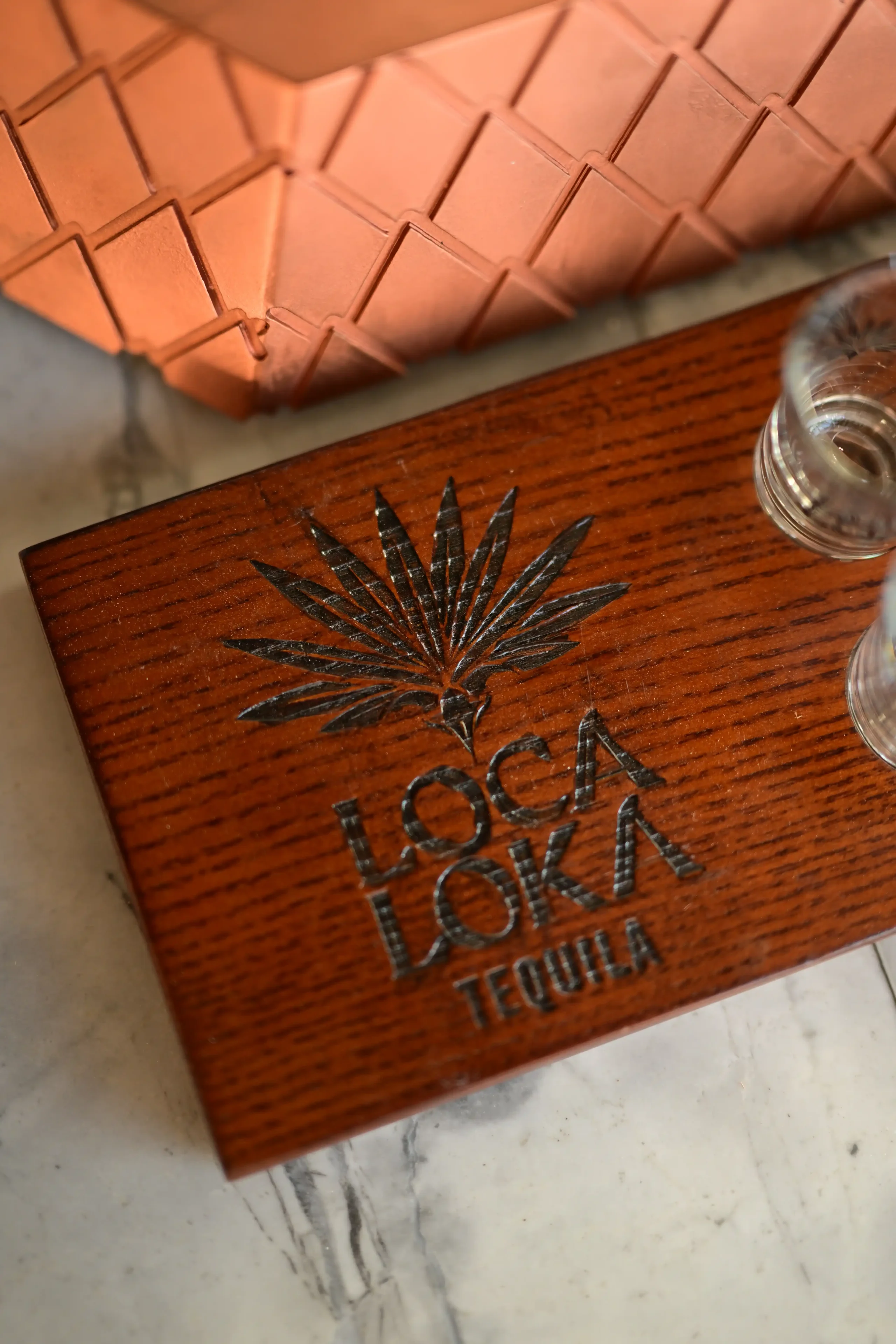 Close-up of a wooden box with a black sunburst logo and the word TEQUILA printed on it, resting on a marble surface with a glass nearby.