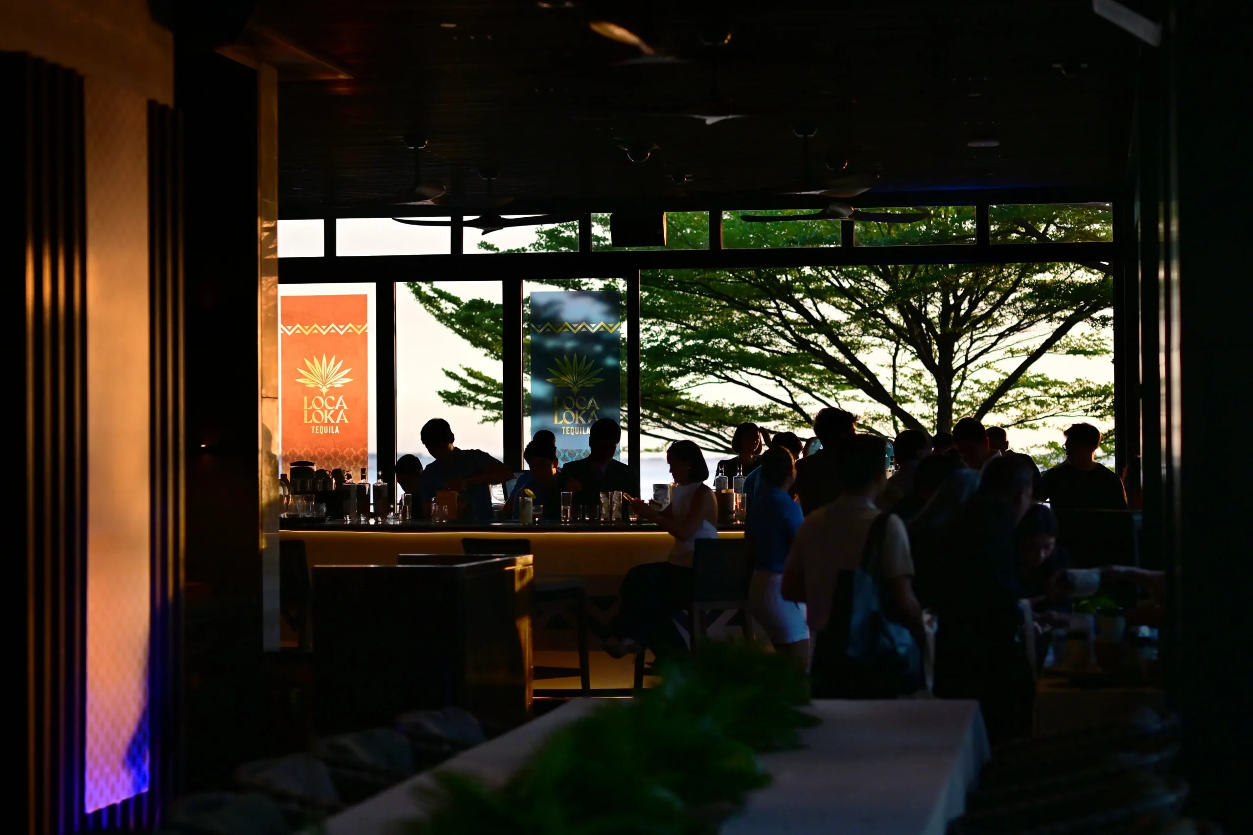 Dimly lit tequila bar with patrons seated at a long counter; outside windows show green trees and sunset, with LOCA LOKA tequila banners visible.