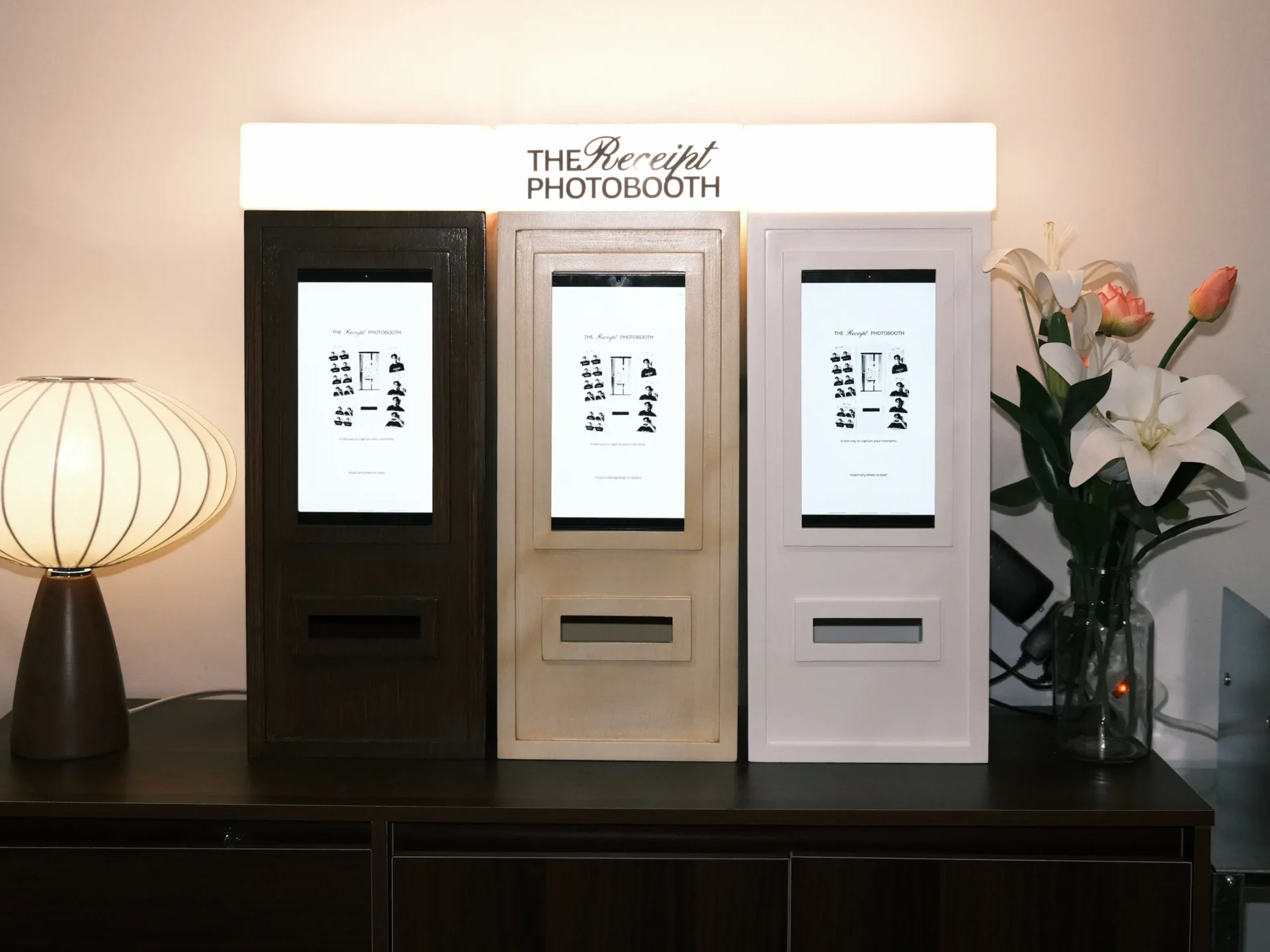 Three photo booths in dark, beige, and light gray finishes stand in a row beneath a sign reading 'The Receipt Photobooth', with a lamp on the left and a vase of lilies on the right.