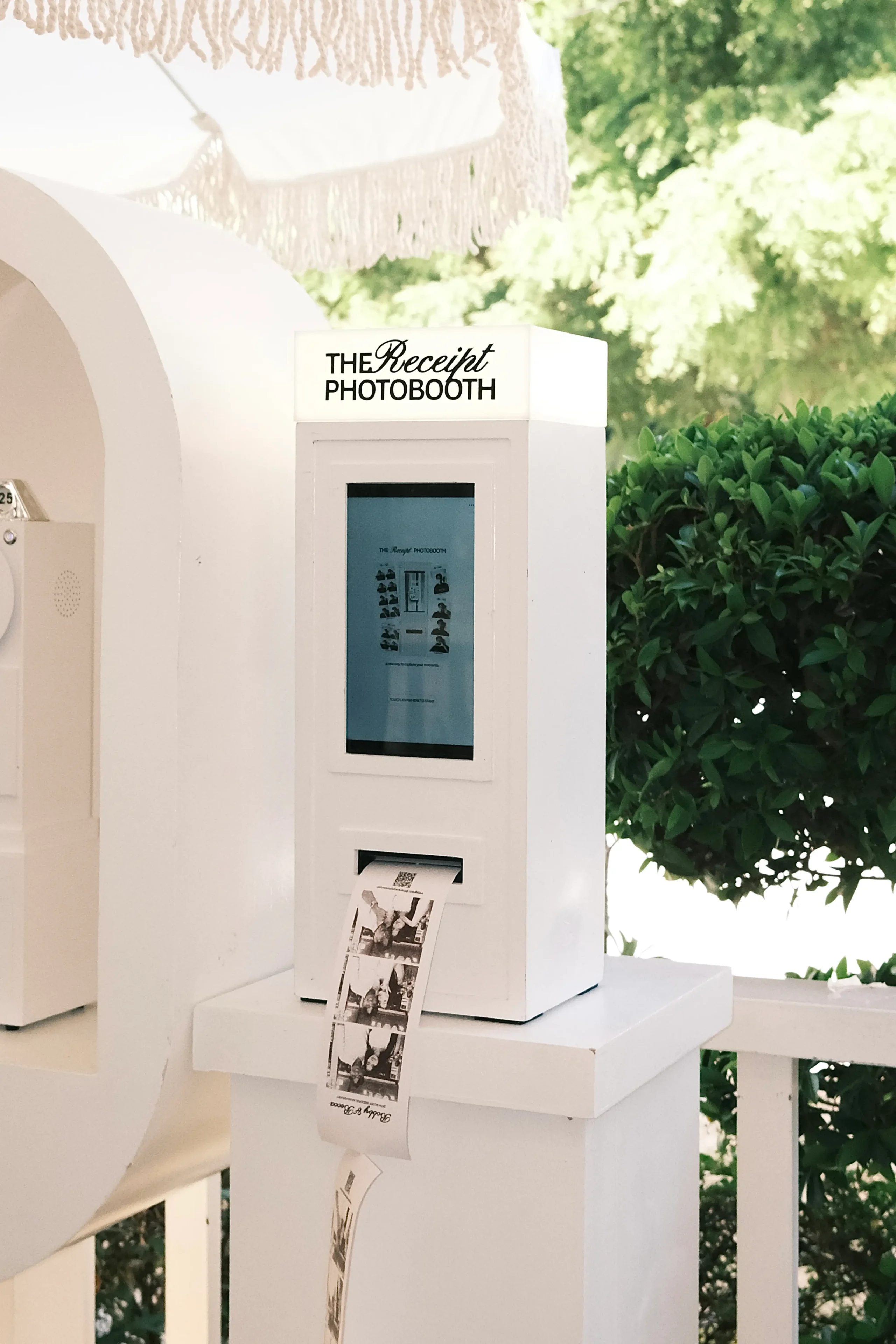 White photo booth labeled 'The Receipt Photobooth' with a digital screen and rolling photo strips, placed outdoors among green hedges.