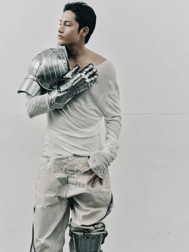 Man in a white outfit with a metallic knight armor gauntlet and shoulder plate on his left arm, looking off to the left.