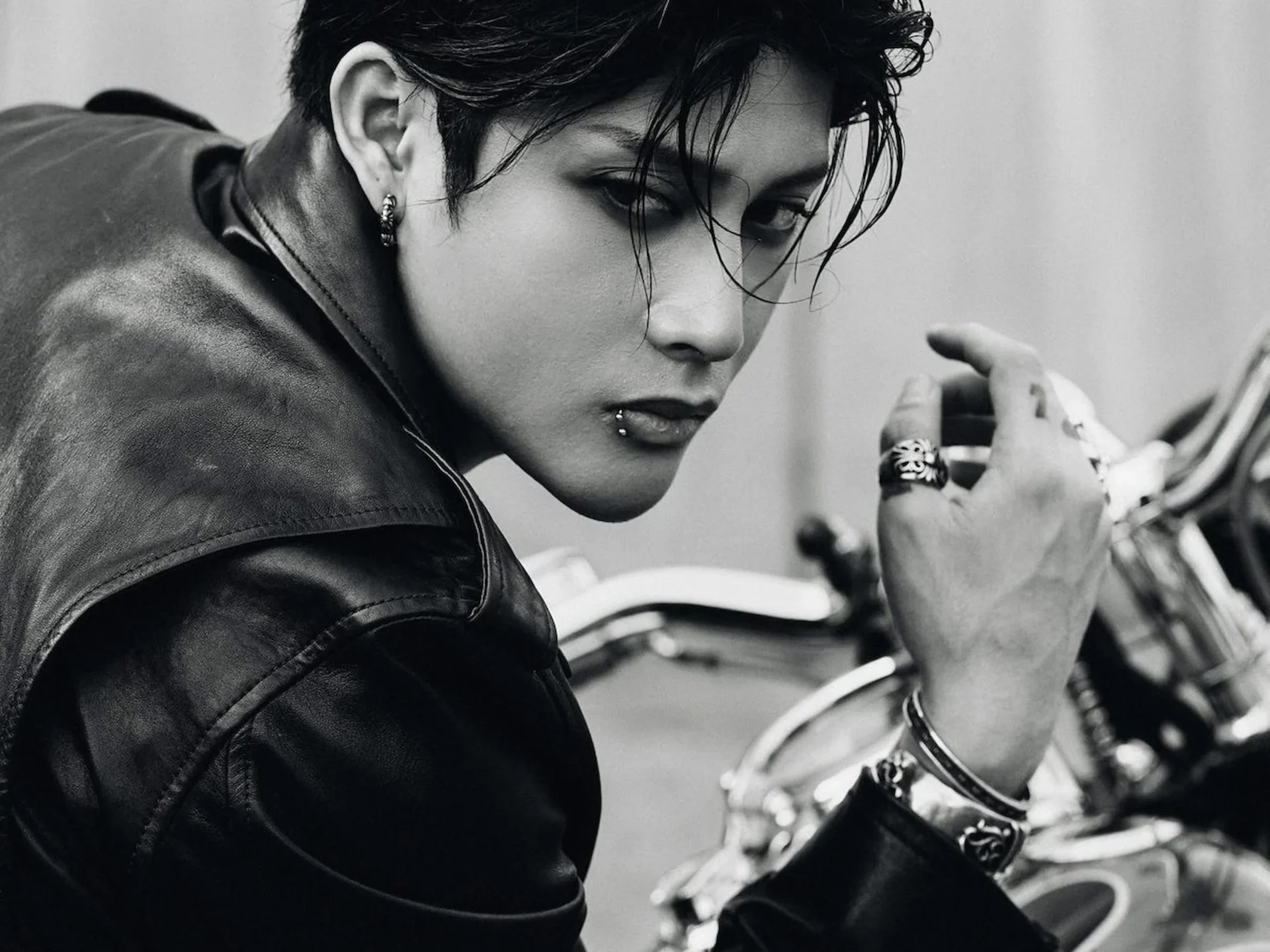 Close-up of a woman in a leather jacket looking downward, while a second person adjusts rings near a motorcycle.