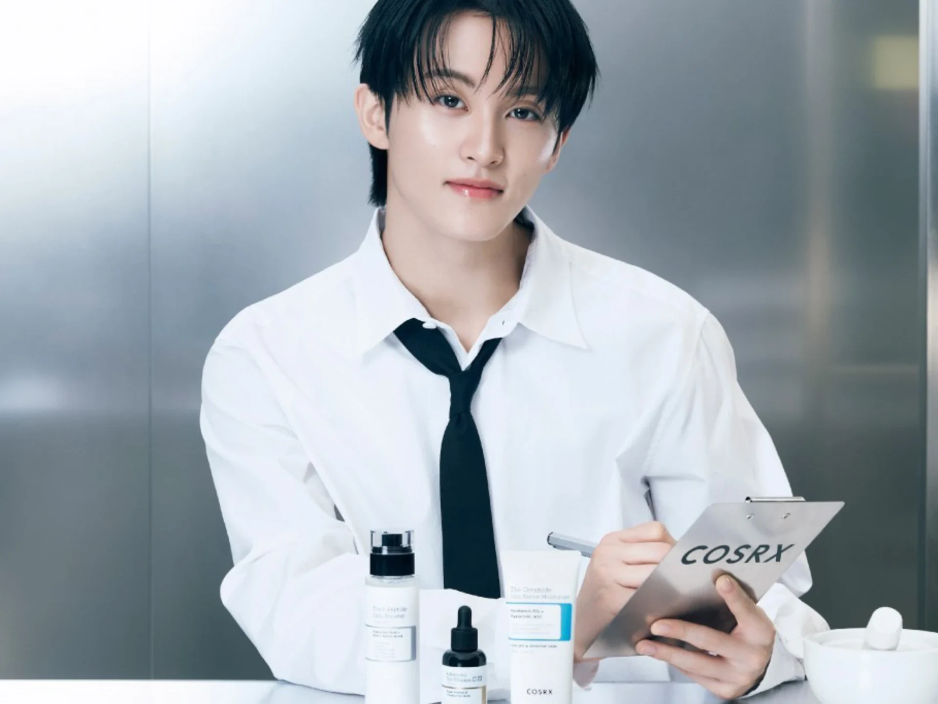 Person in a white shirt and black tie holding a COSRX brochure in a sleek, stainless-steel setting with product bottles nearby and a clipboard visible.