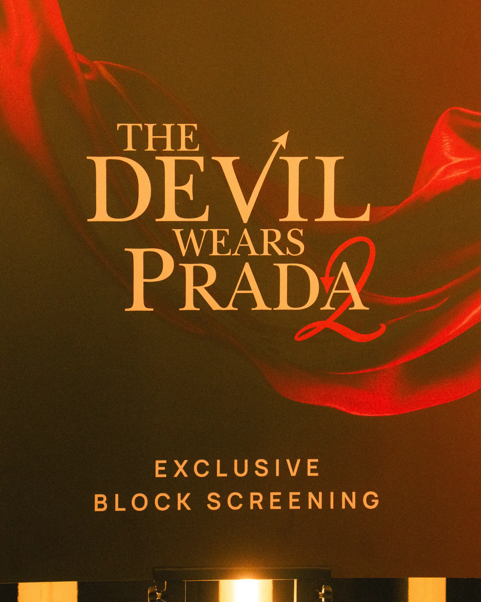 Poster for an exclusive block screening of The Devil Wears Prada on a dark background with red scarf motif and white title text