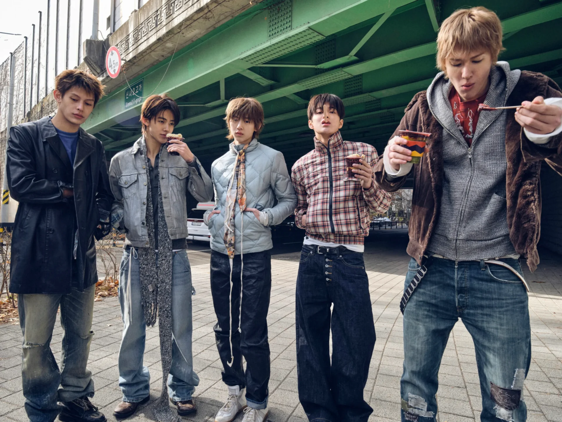 Five young men stand under a green elevated railway, eating snacks from cups under the structure.