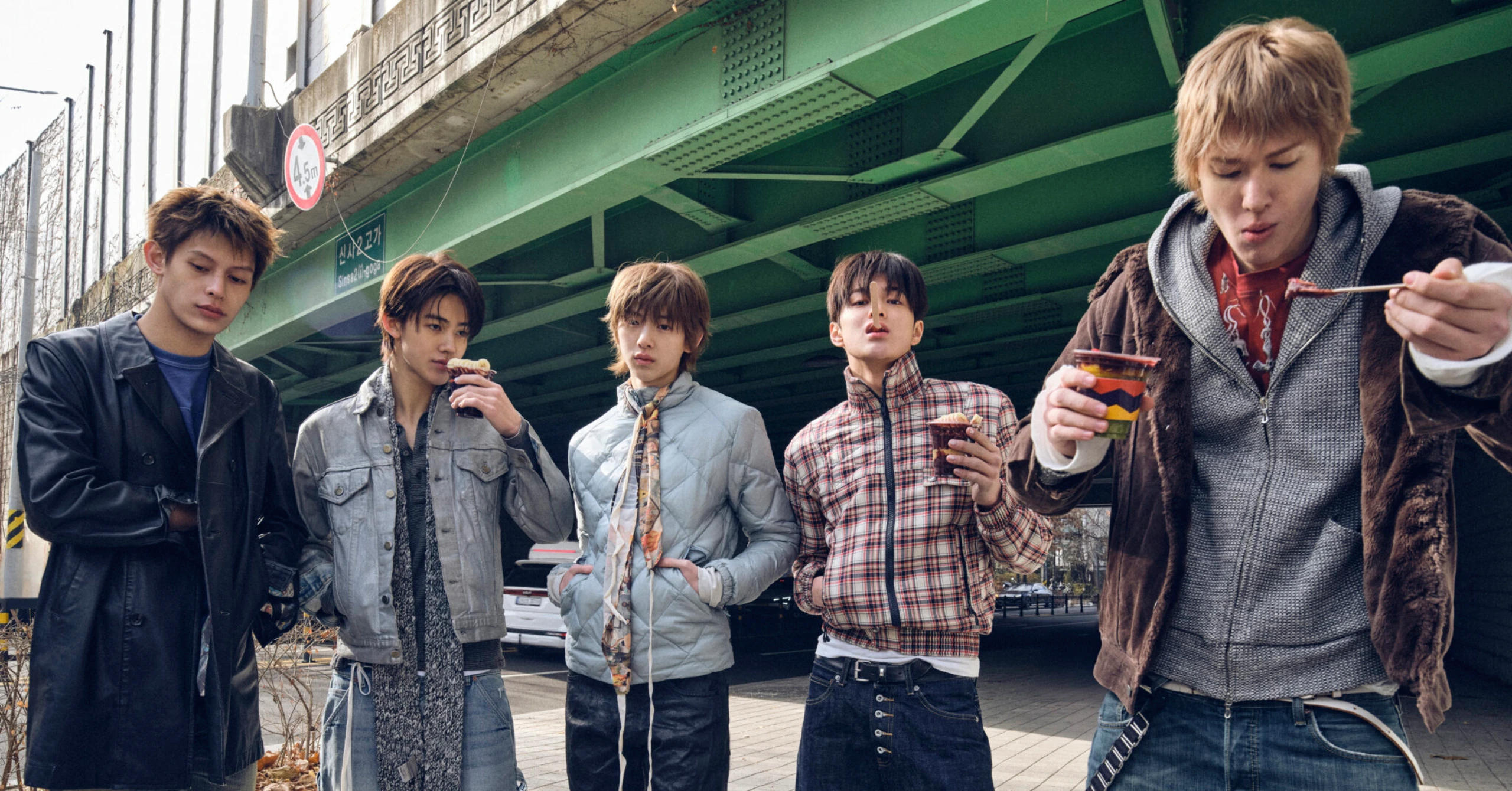 Five young men stand under a green elevated railway, eating snacks from cups under the structure.