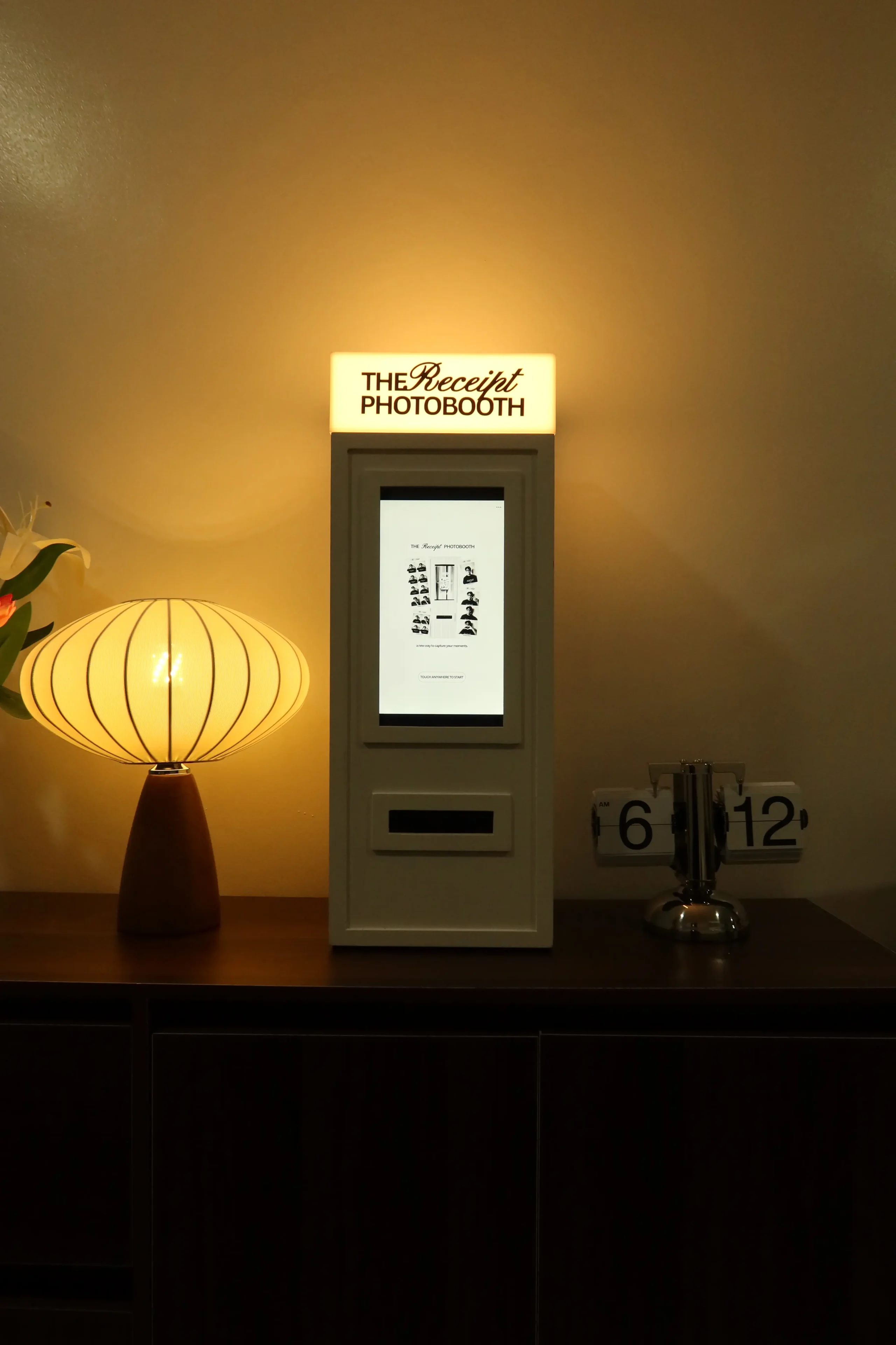 Photo booth labeled 'The Receipt Photobooth' on a counter with a digital screen, beside a warm lampshade lamp.