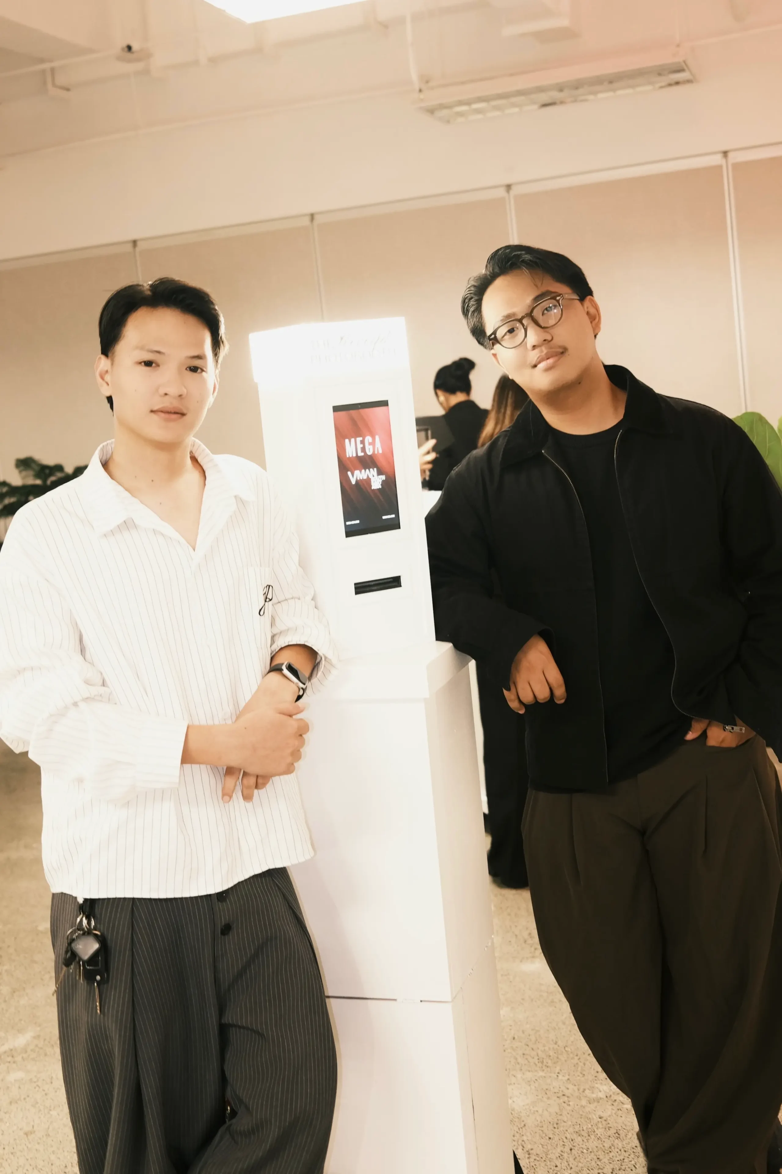Two men pose beside a white display stand in a bright indoor space; left man wears a white striped shirt and a watch, right man wears a black jacket and glasses.